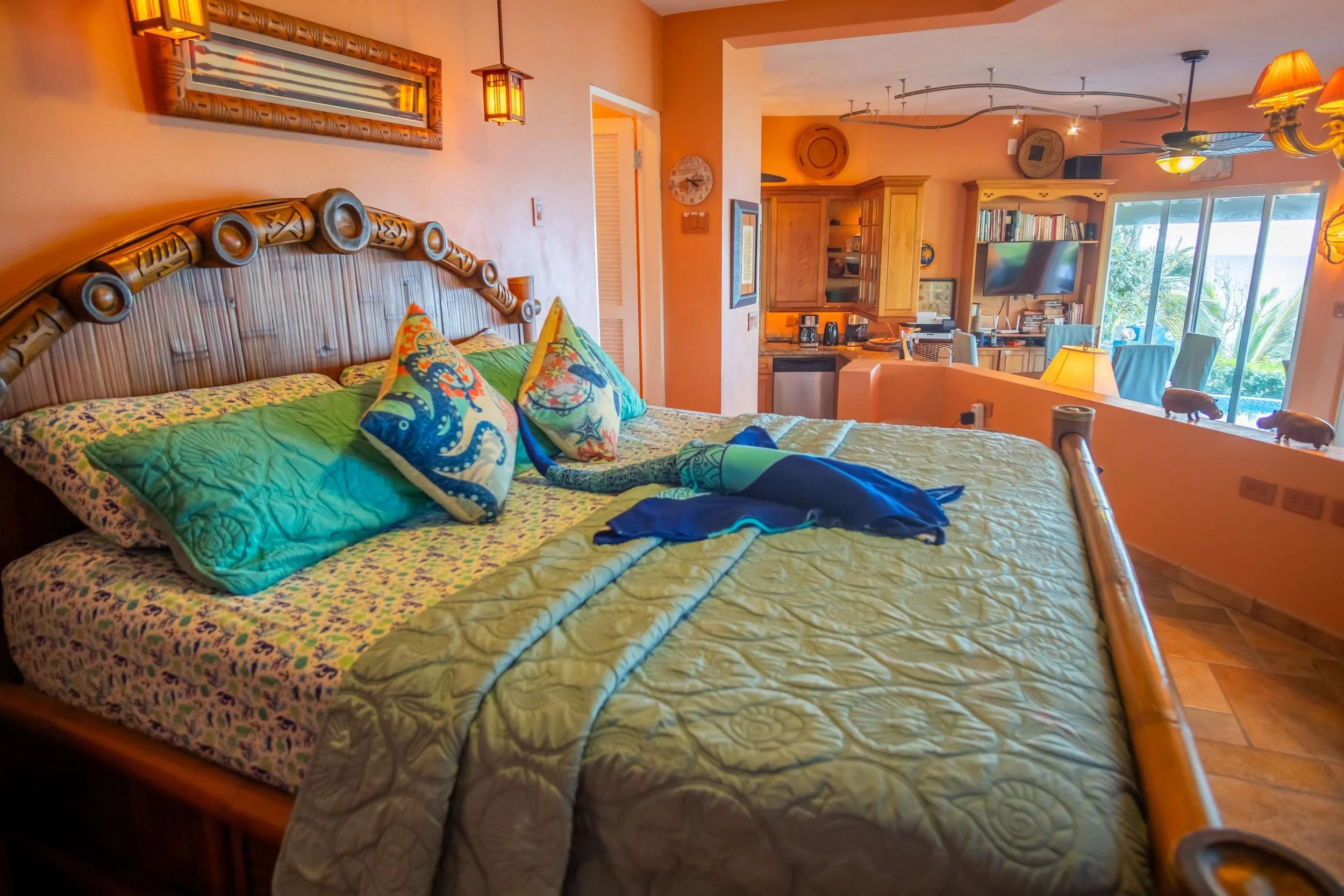 A tropical-themed hotel room with a large bed, colorful pillows, and an ocean scene painting on the wall; the background features a kitchenette and a dining area with sliding glass doors revealing a lush outdoor view.
