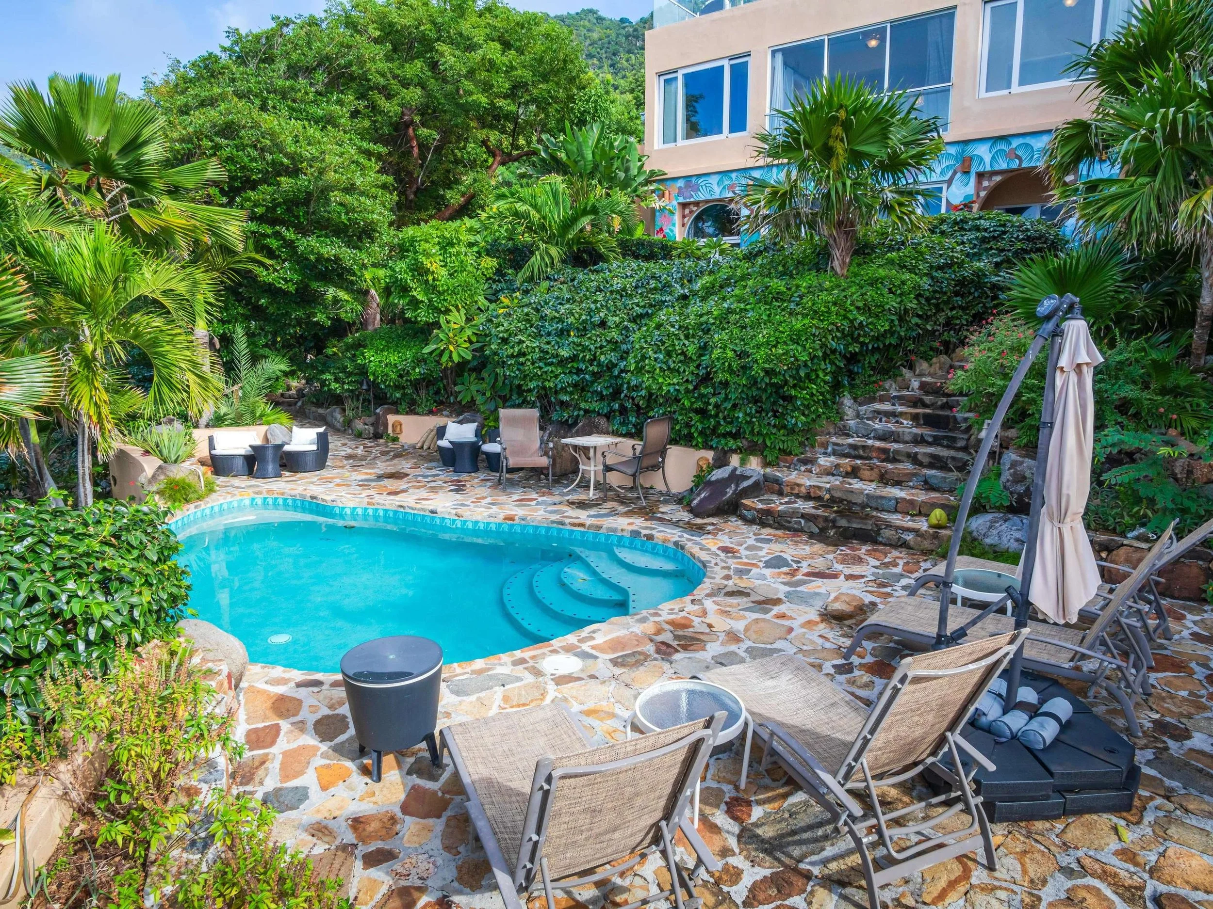 Small swimming pool surrounded by stone patio, lush green tropical plants, outdoor chairs, umbrellas, and a building in the background.