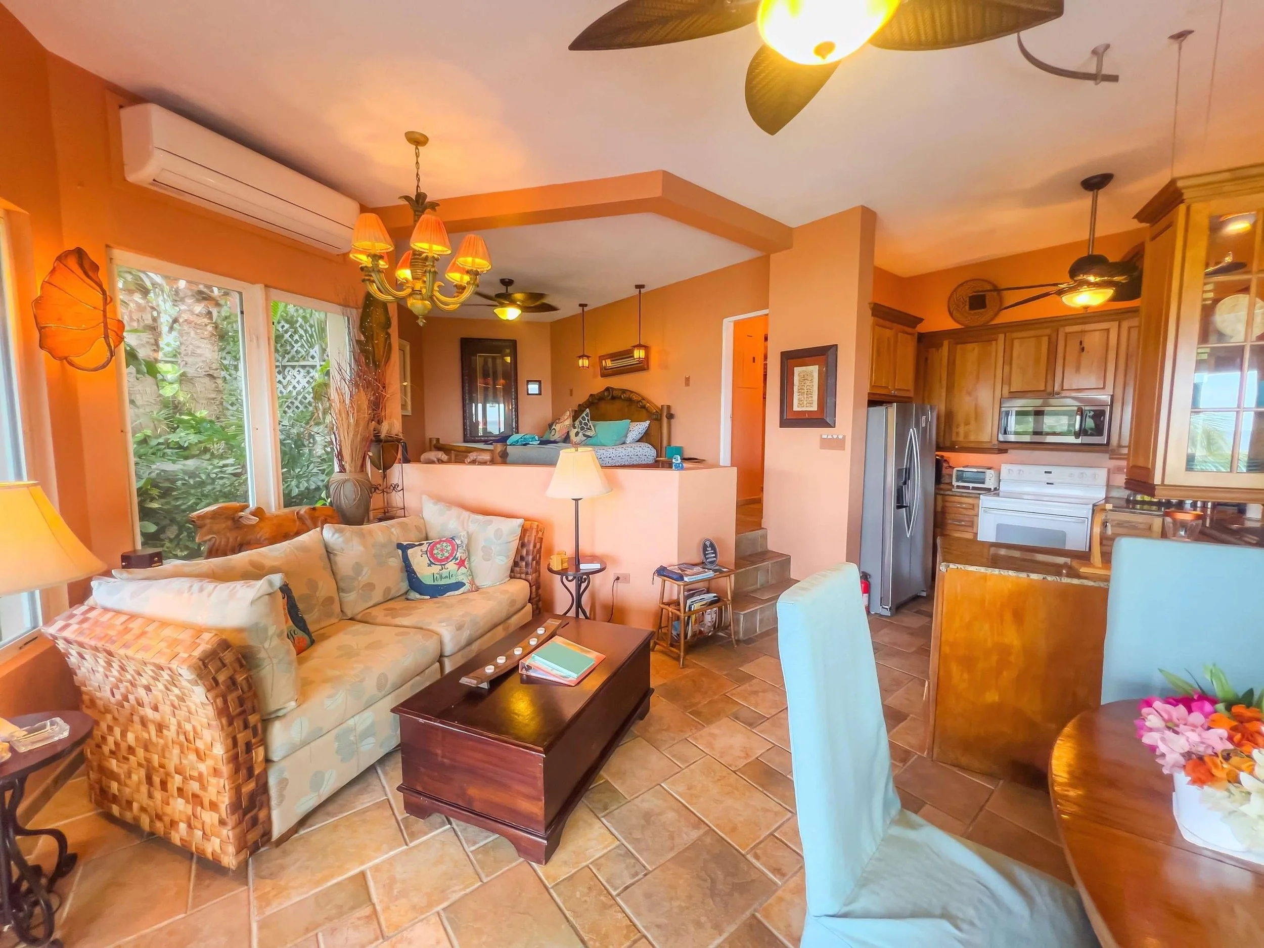 A cozy living room with a beige sofa with patterned pillows, a dark wooden coffee table, and a light blue chair. There's a dining table with a flower arrangement and a kitchen with wooden cabinets, a stainless steel refrigerator, and white appliances