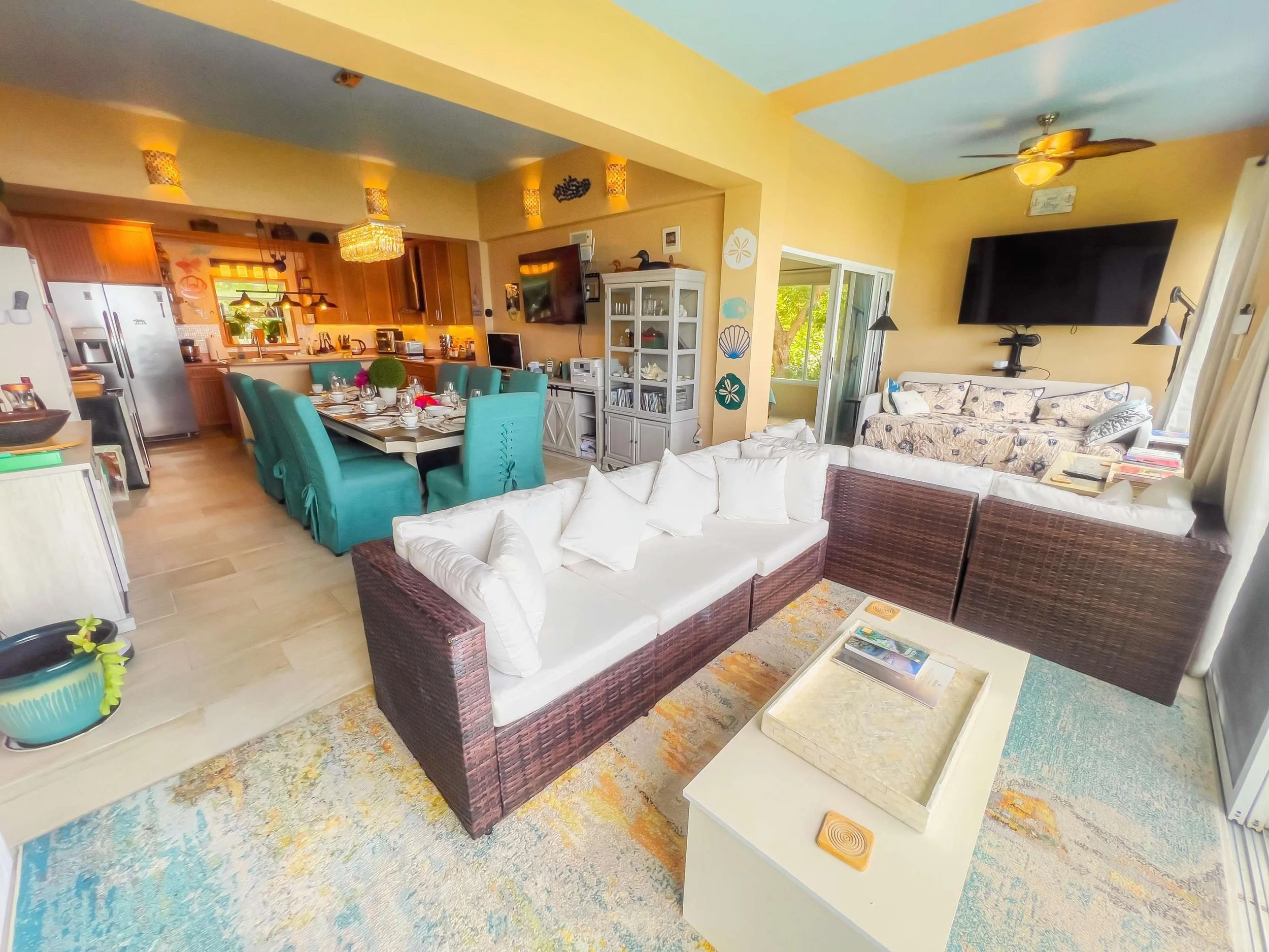 Open-concept living and dining area with white and brown sofas, a white coffee table, dining table with teal chairs, and a kitchen with wooden cabinets and stainless steel appliances. Large windows and sliding glass door lead to a balcony, with ceili