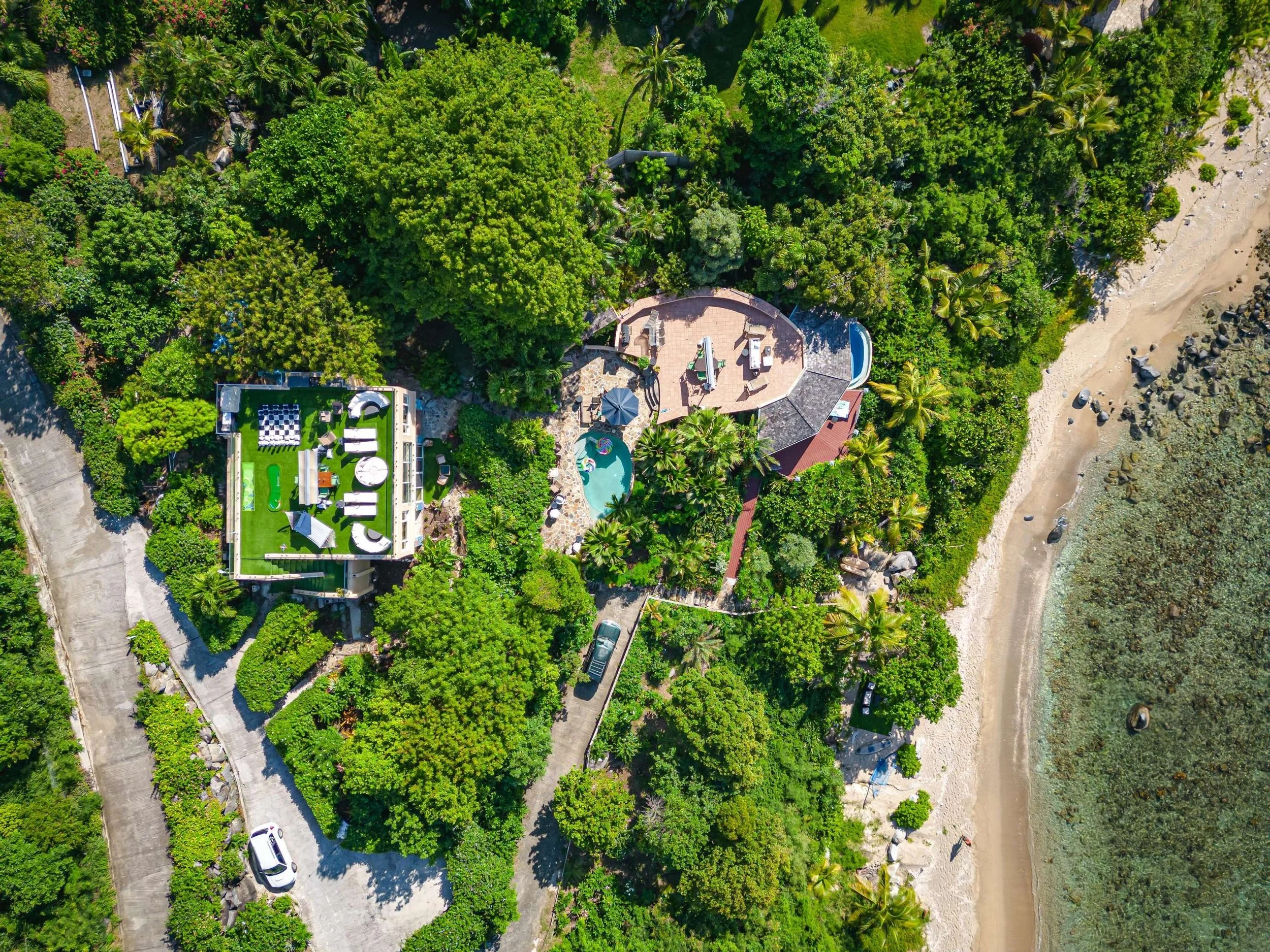 Aerial view of a lush green outdoor area with two houses, one with a green roof and modern design, and the other with a brown roof, surrounded by dense trees and shrubs. There is a beach with rocks on the right side of the image.