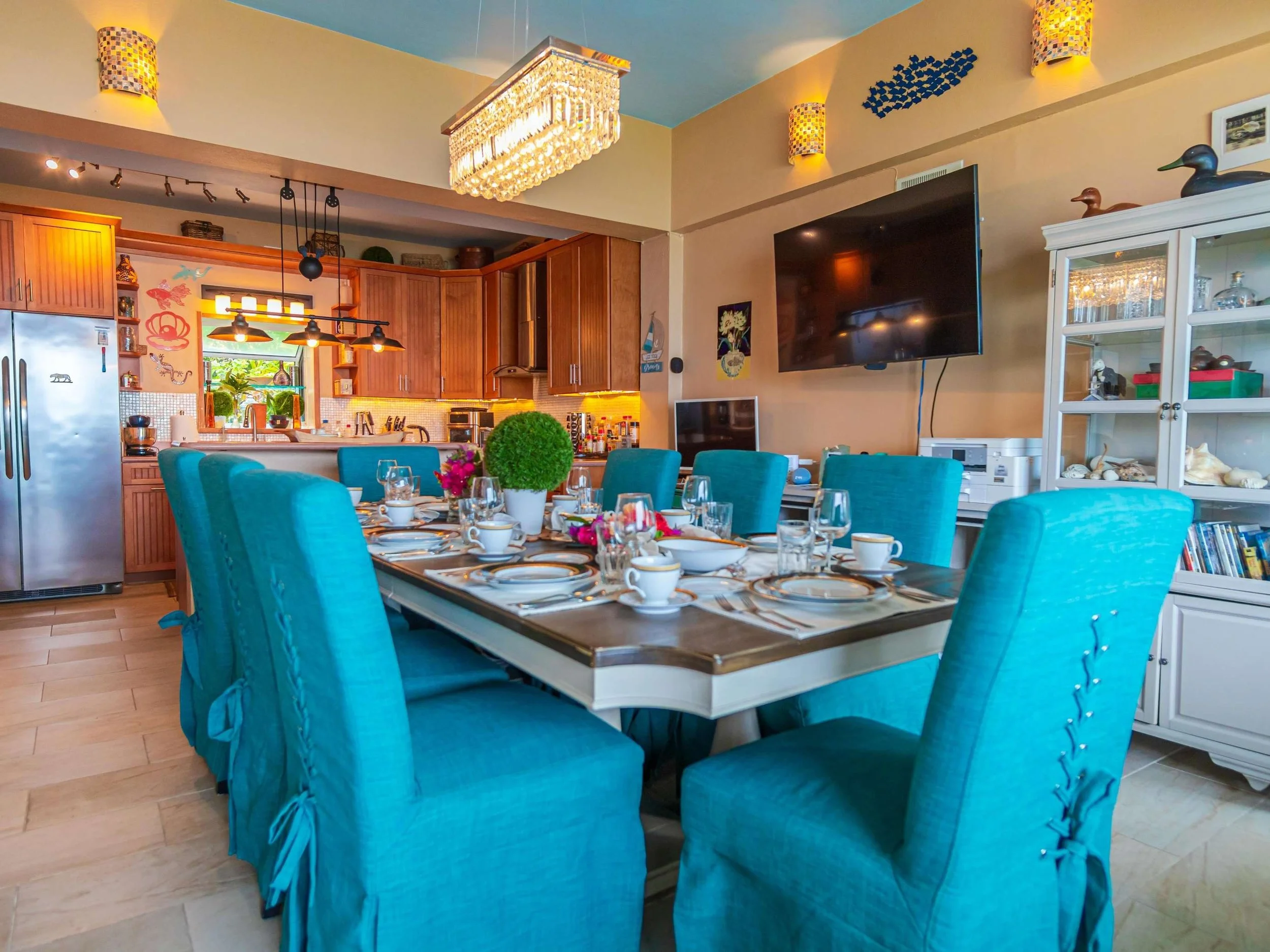 A dining room with a large table set for a meal, surrounded by eight teal upholstered chairs. The room opens into a kitchen with wooden cabinets and stainless steel appliances. Decor includes a large chandelier, wall-mounted fish artwork, and nautica