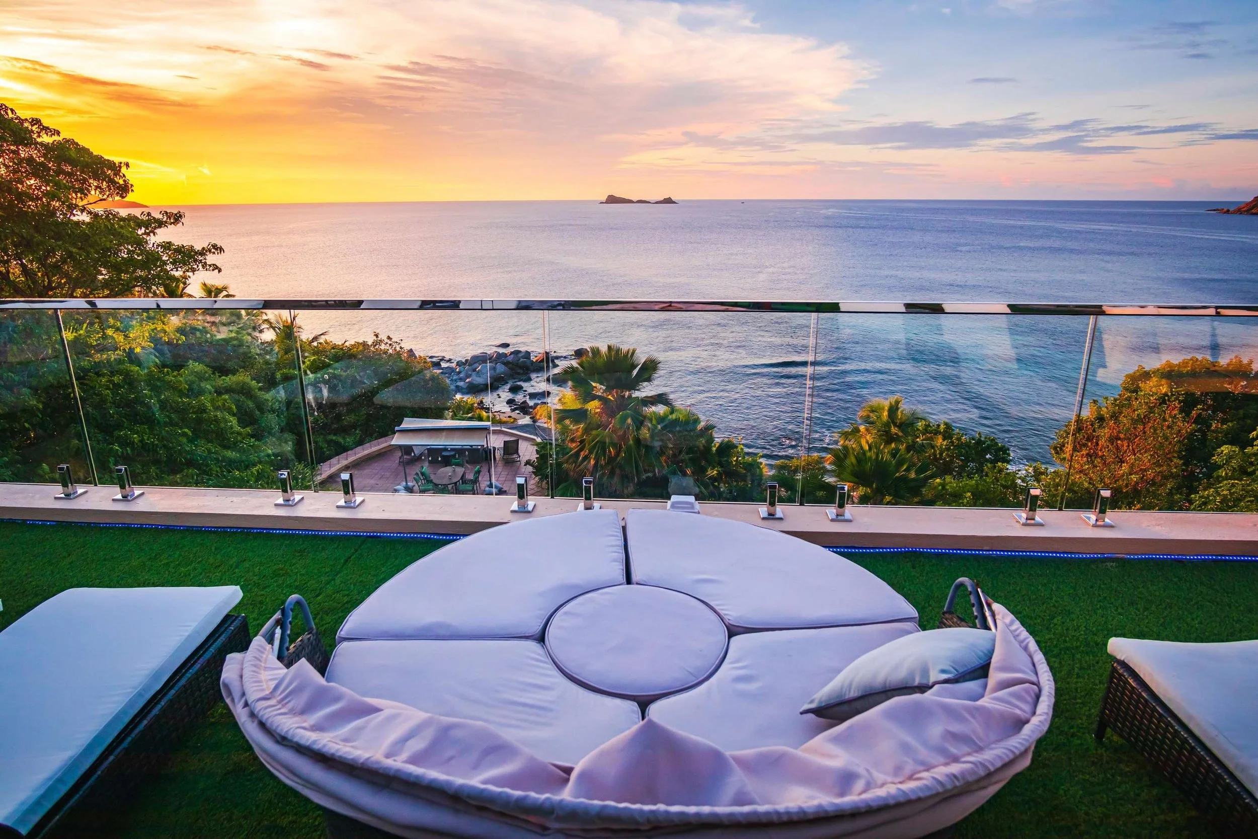Luxury balcony with outdoor seating, overlooking a sunset over the ocean with trees and rocks below.