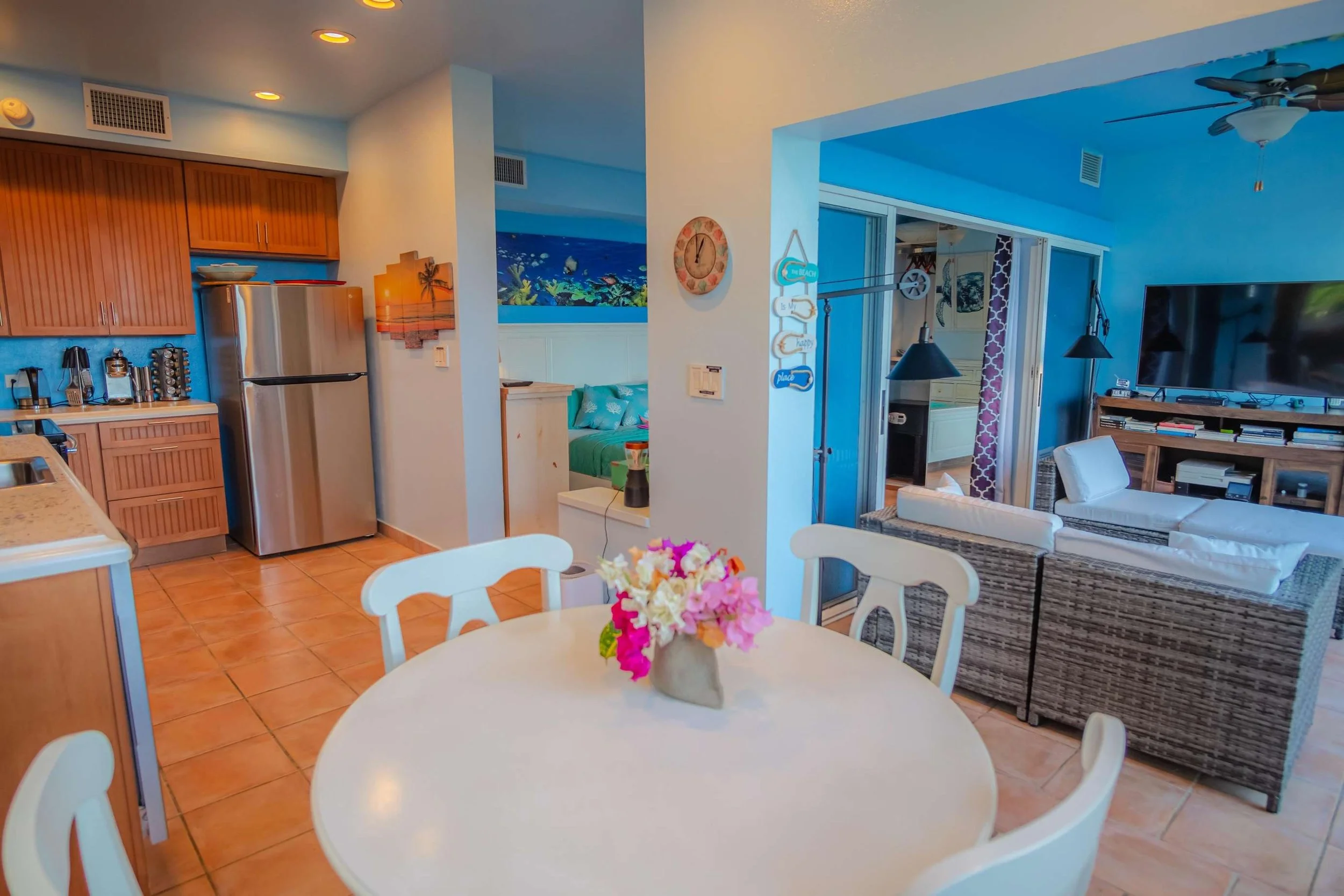 A dining area with a round table and a vase of colorful flowers, located in a kitchen that opens up to a living room, with visible kitchen appliances, a wall with tropical artwork, and a bedroom in the background with vibrant blue walls and seaside d