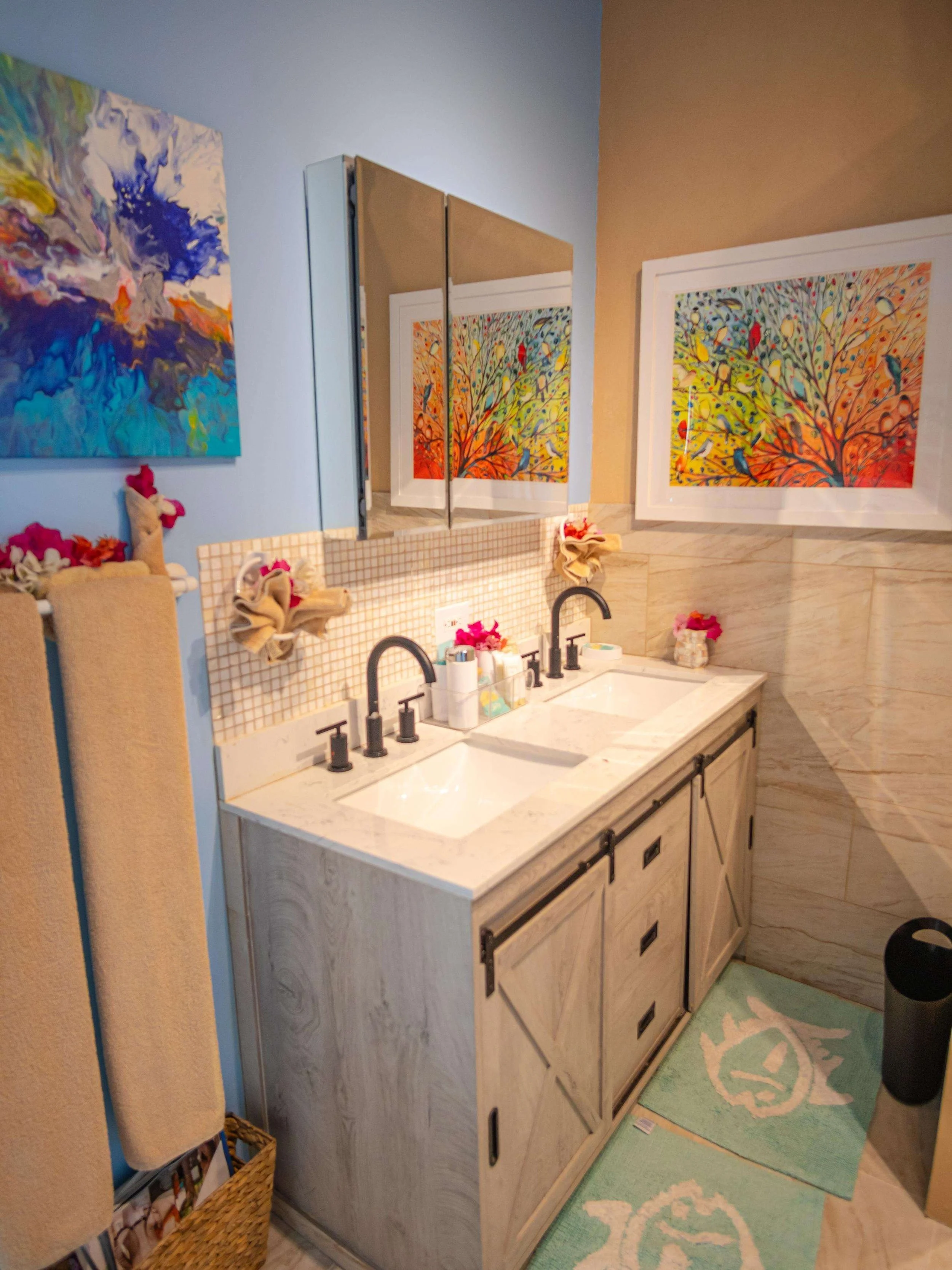 Bathroom with dual sinks, beige wooden cabinet, colorful artwork on the walls, mirror, towels, and decor.