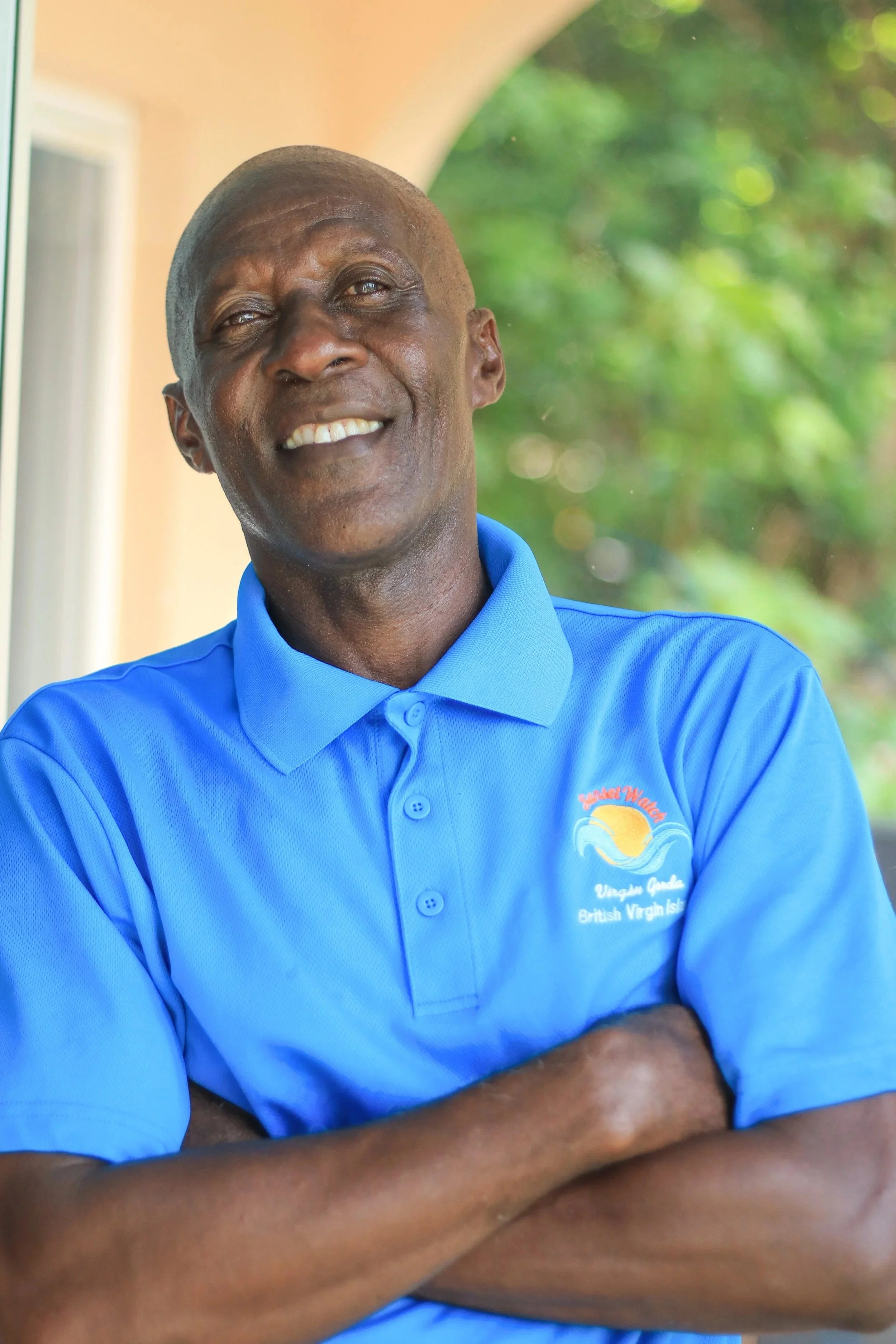 A smiling man with crossed arms, wearing a bright blue polo shirt with a logo, standing outside in front of a green leafy background.