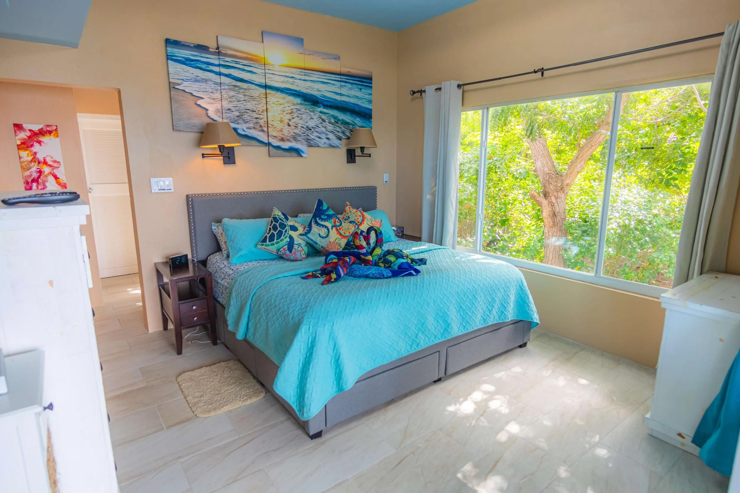 Bedroom with a large window showing green trees outside, a bed with a light blue quilt and colorful pillows, a nightstand, and a wall art depicting the beach at sunset.