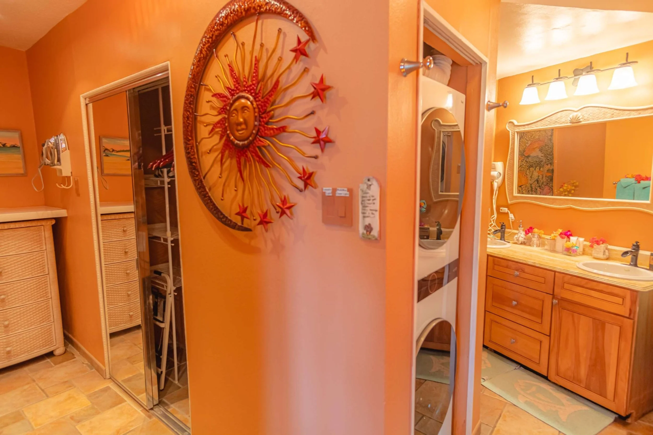Interior view of a bathroom with double sinks, a large mirror, decorated with flowers, and a small washer and dryer unit. The walls are painted orange, and there is a sun-themed wall decoration with a face and rays on the wall.