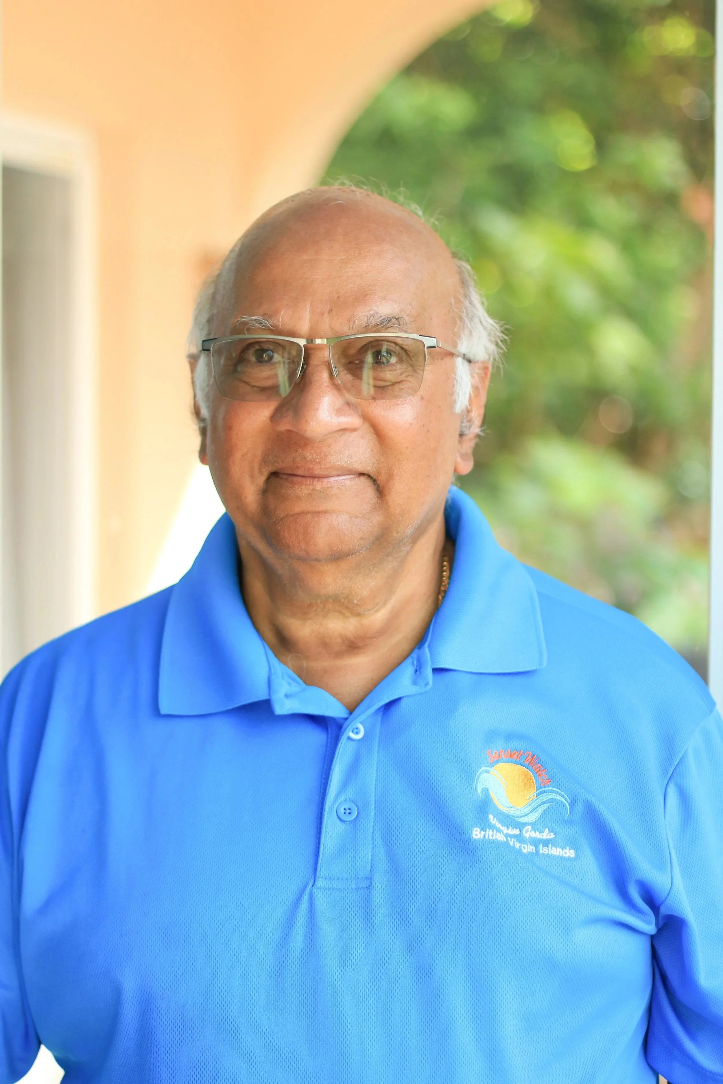 A man with glasses and a blue polo shirt standing outdoors, with trees in the background.