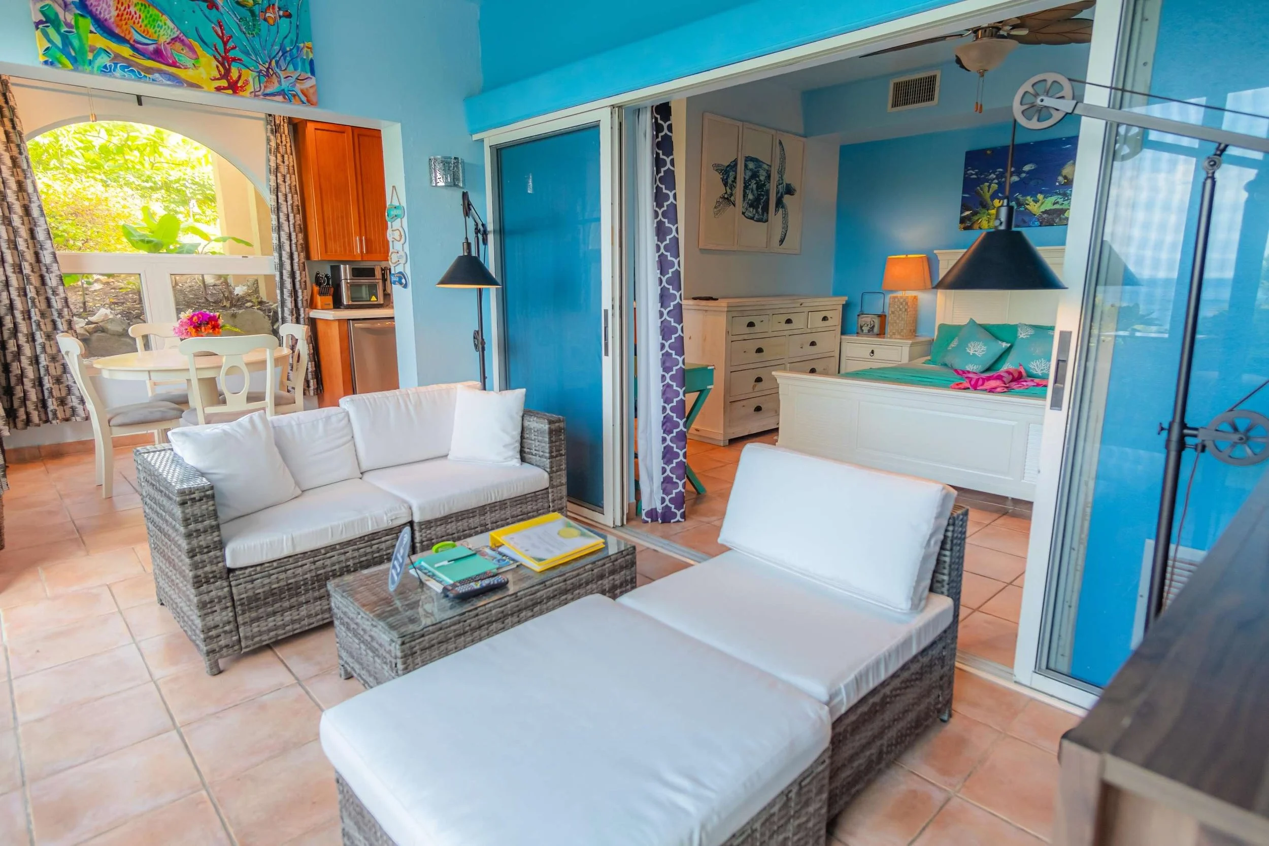 Living room with wicker furniture, a white sofa, and a chaise lounge, opening into a bedroom with a white bed, two lamps, artwork, and a dresser, adjacent to a dining area with a round table and chairs, decorated with colorful paintings and curtains,