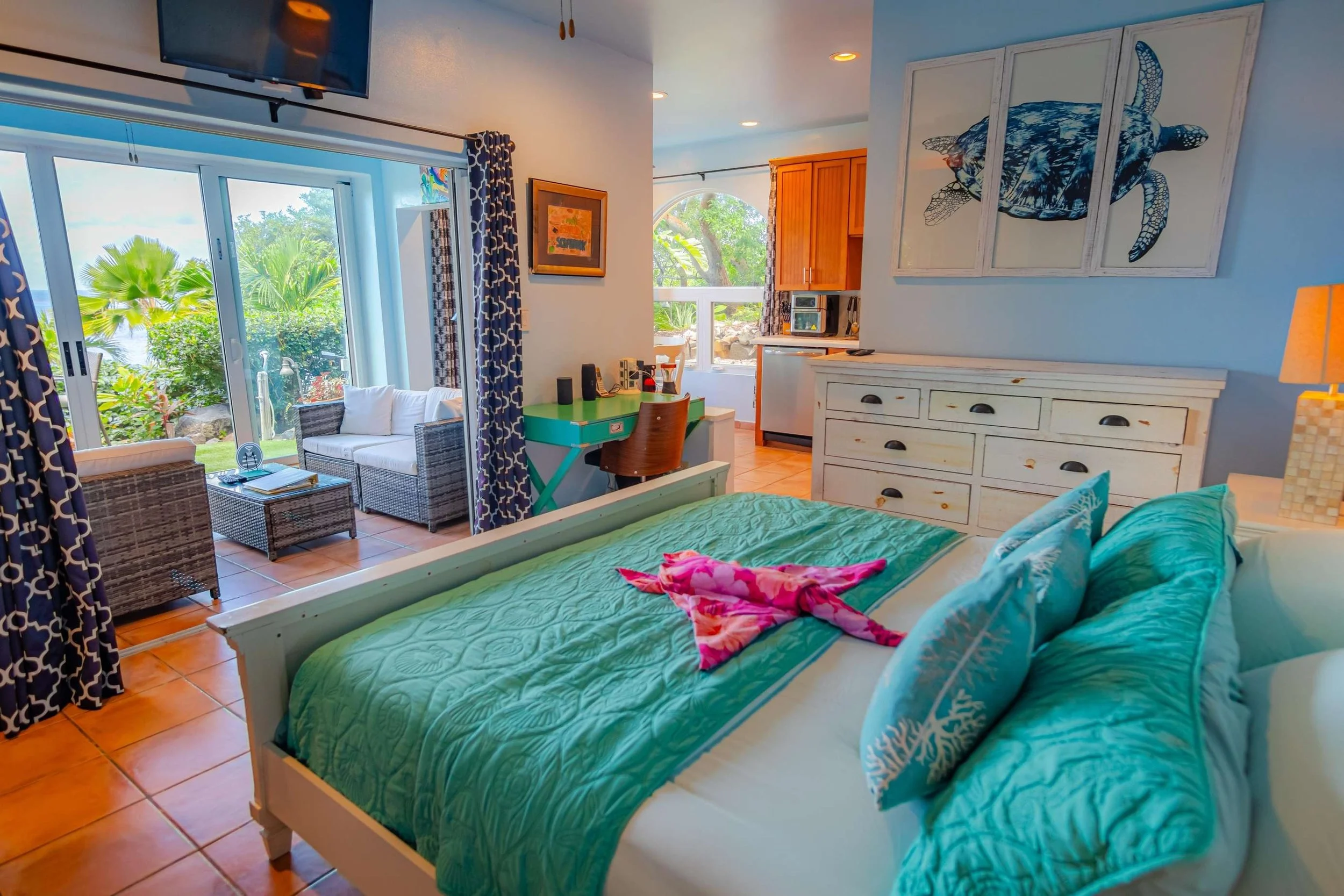 A cozy bedroom with a teal bedspread and pillows, a white dresser, and tropical decor. A sliding glass door opens to a patio with outdoor seating and lush greenery outside.