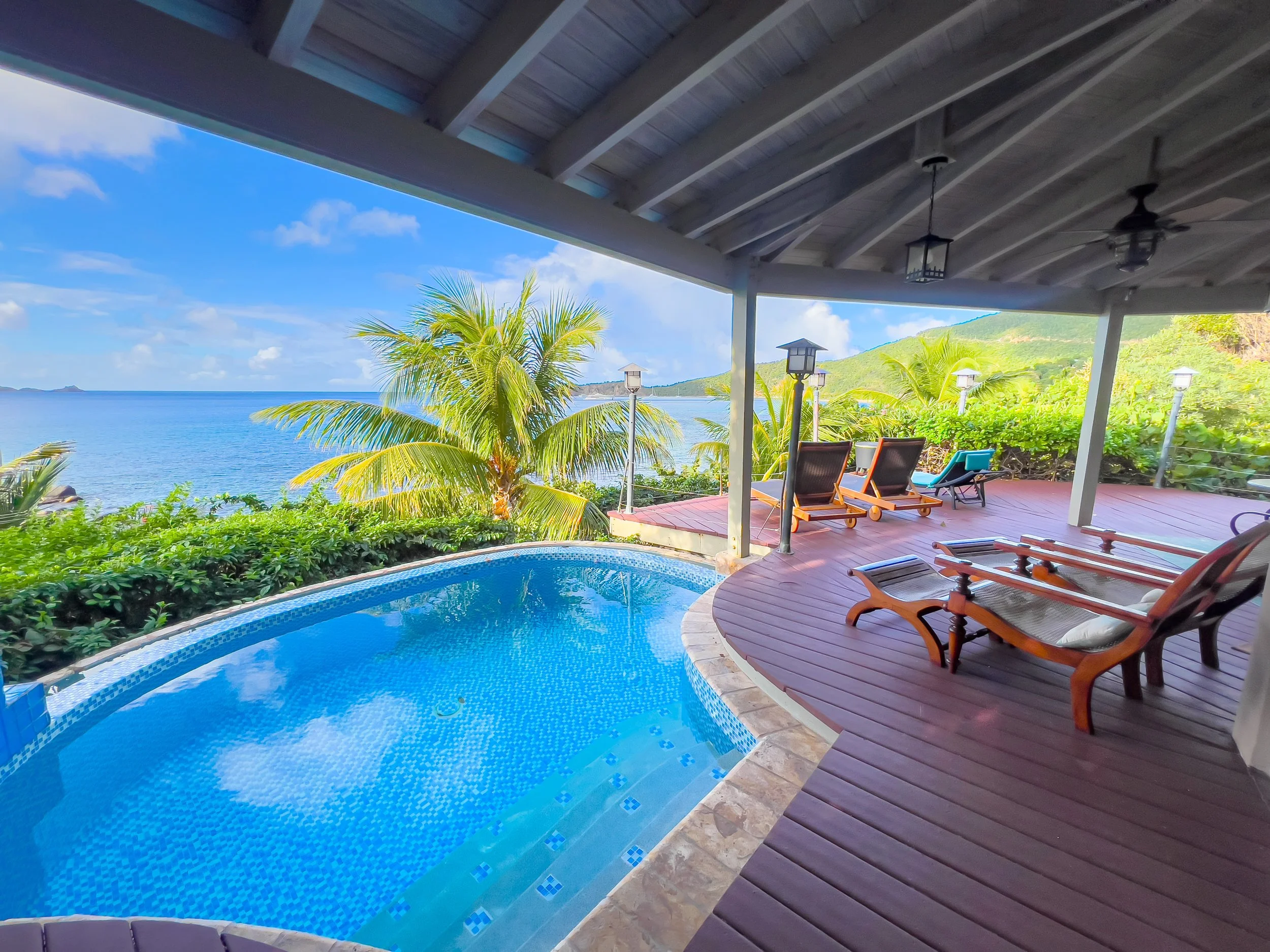 Covered patio with lounge chairs overlooking a tropical beach, swimming pool, and palm trees under a partly cloudy sky.