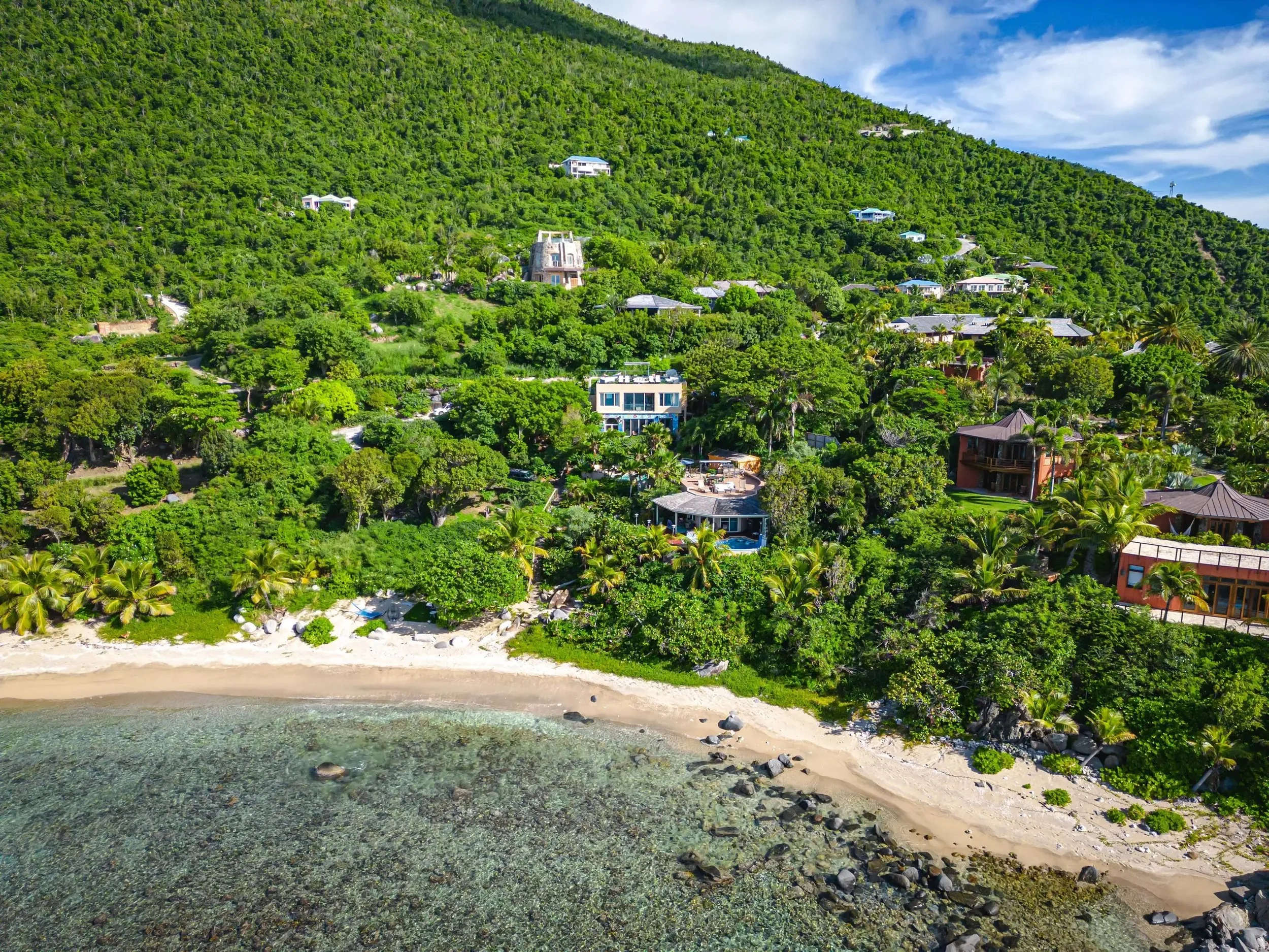 Aerial view of a coastal hillside with multiple houses surrounded by lush green trees and vegetation, with a sandy beach and clear water in the foreground.
