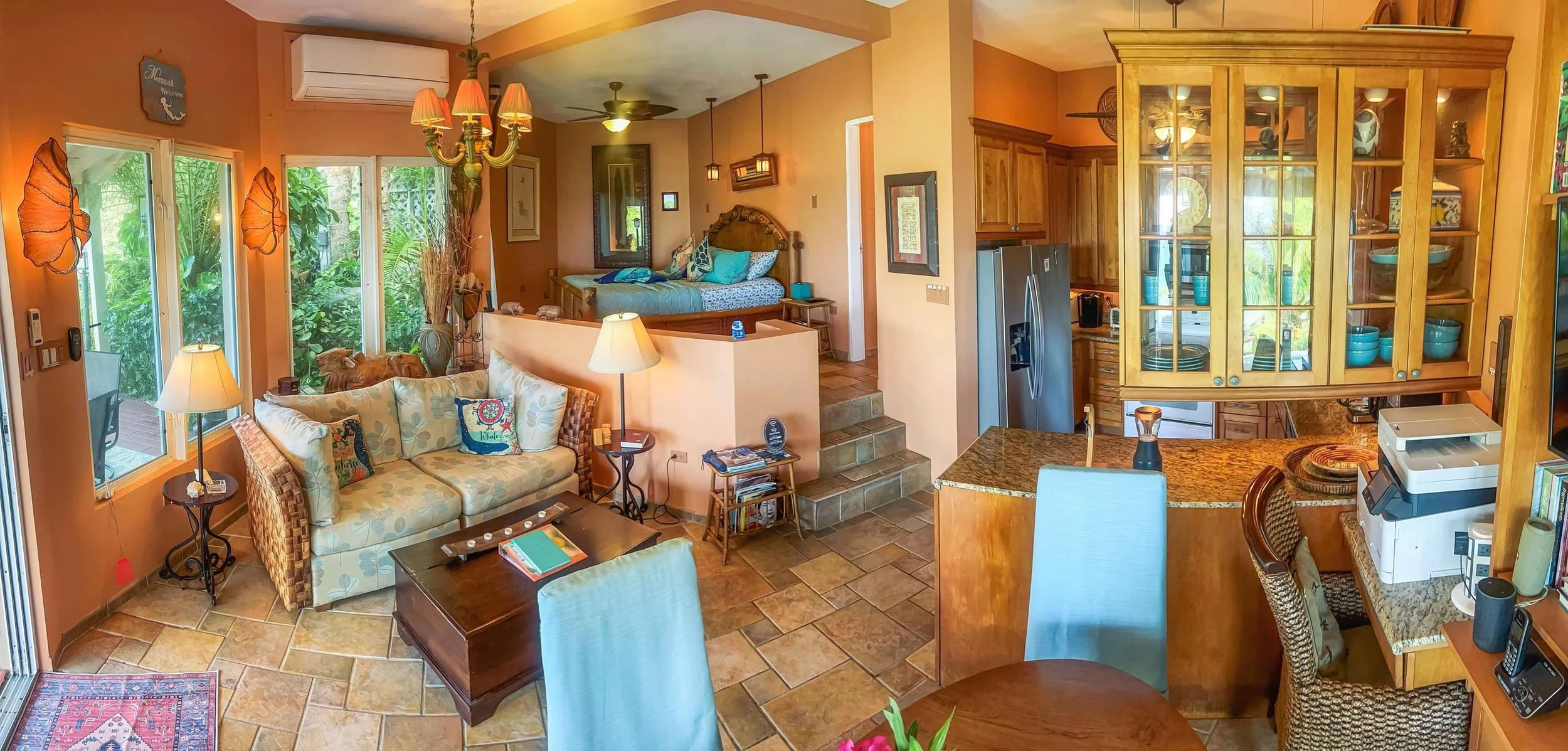 A cozy open-concept living room and kitchen area with large windows showing greenery outside. The living room features a beige sofa adorned with colorful pillows, a coffee table, and a side table with a lamp. The kitchen has wooden cabinetry, a grani