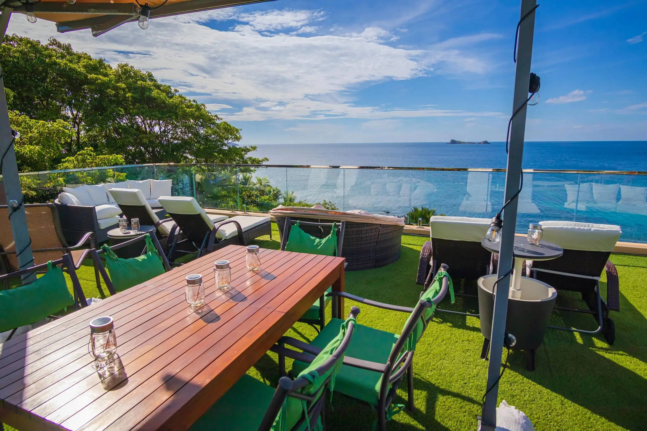 An outdoor rooftop lounge with a view of the ocean, featuring a wooden dining table with mason jar glasses, green cushioned chairs, and lounge seating. A clear glass railing surrounds the area with trees and the sea in the background, and the sky is 