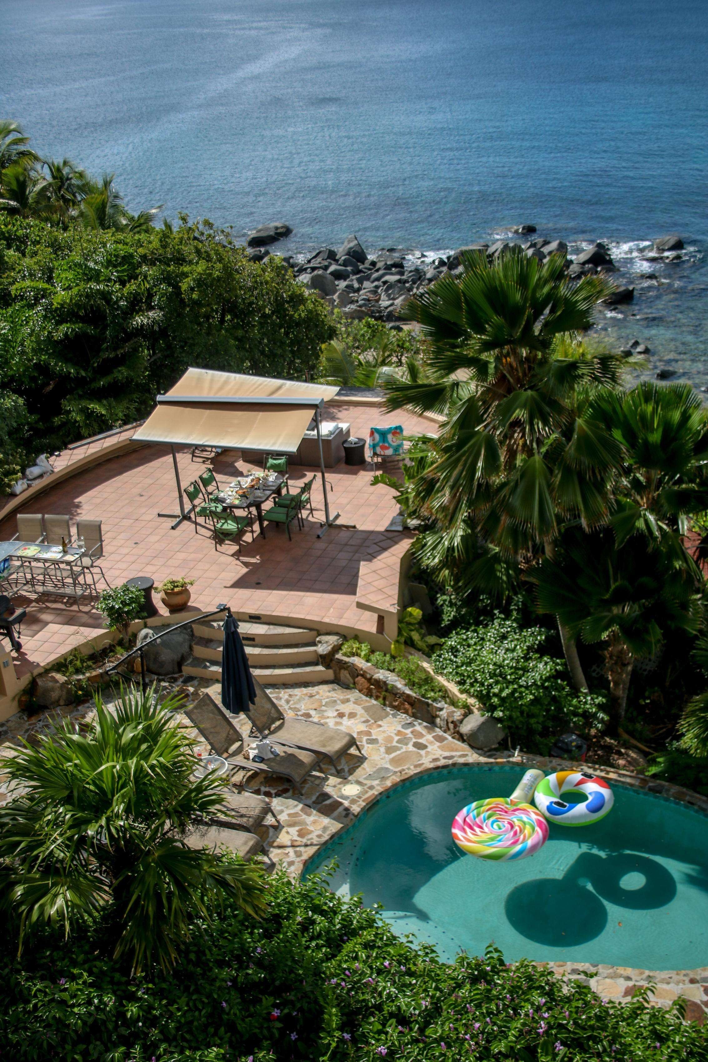 A backyard with a swimming pool, pool floats, lounge chairs, a patio with a dining area, lush green tropical trees, and a view of the ocean with rocks.