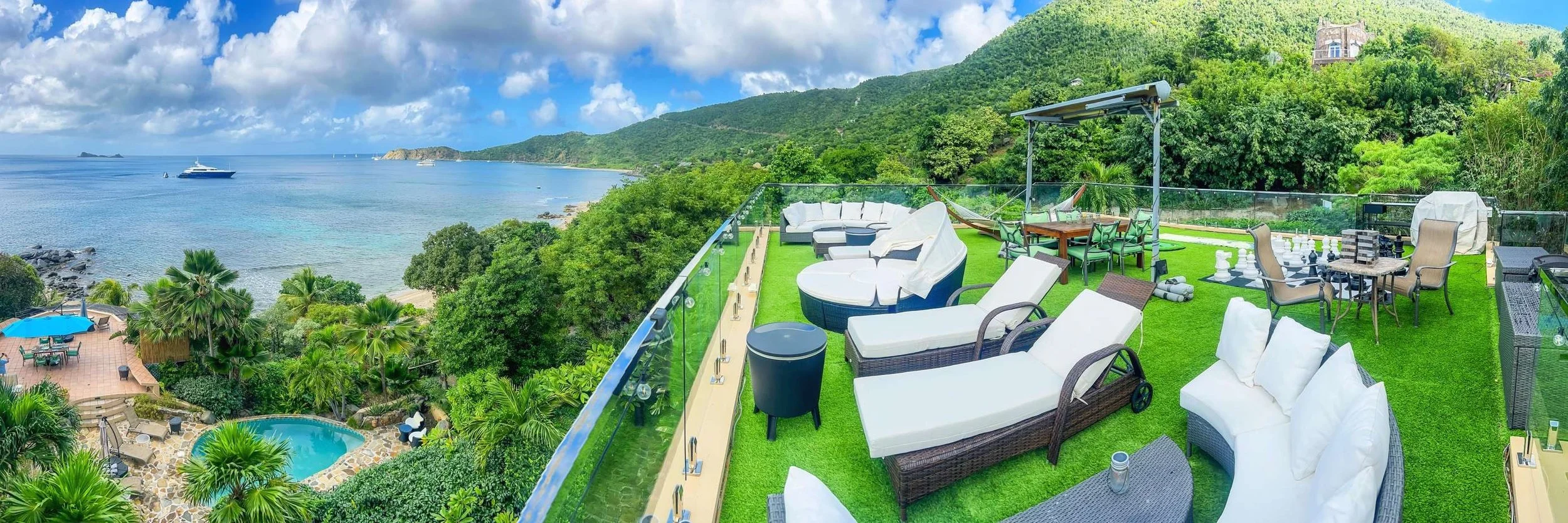 A rooftop terrace with outdoor furniture, including lounge chairs, a dining table with chairs, a hammock, and a chess set, overlooking a coastline with a boat in the sea, lush green trees, and hills in the background.