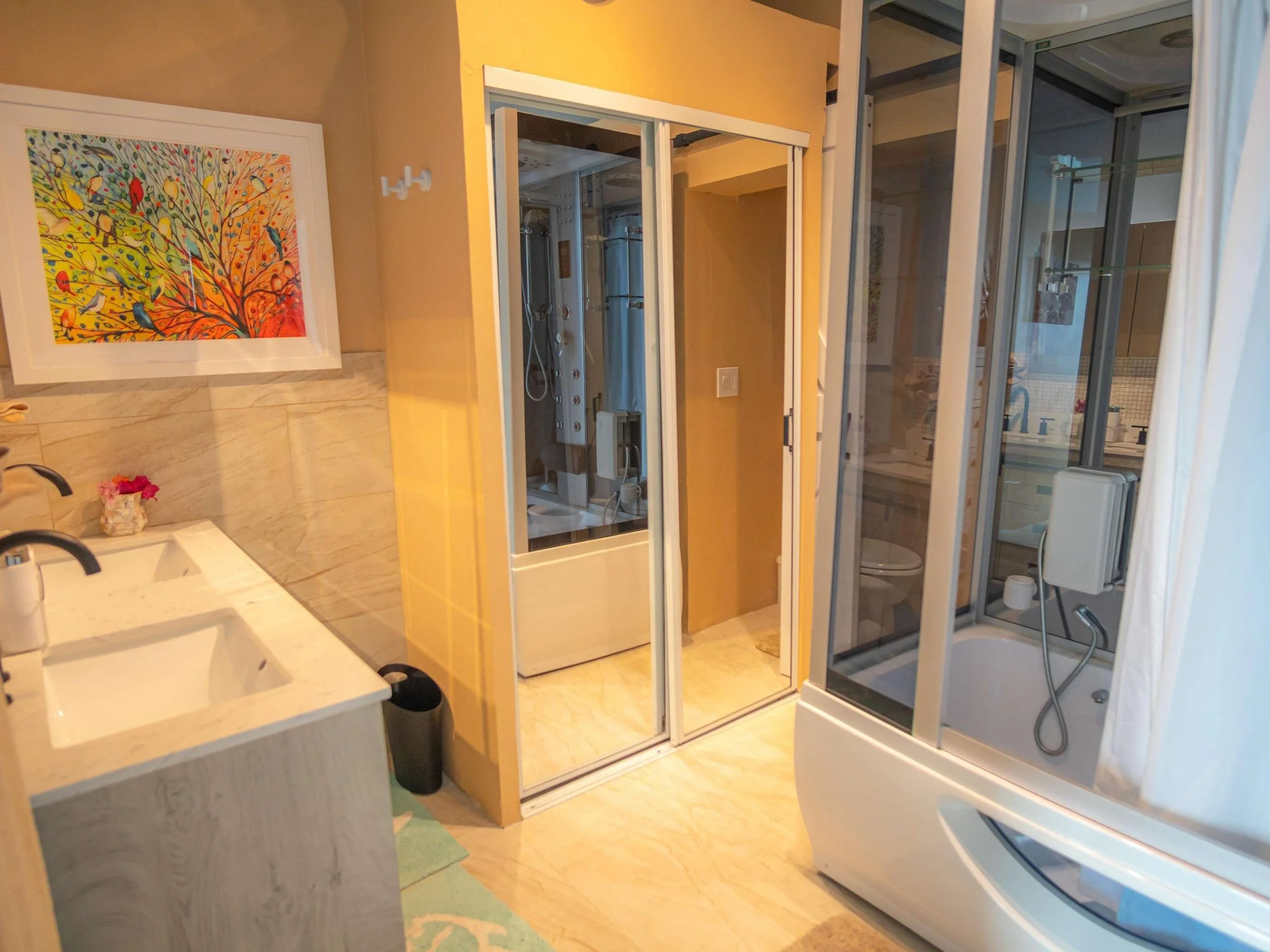 A bathroom with a double sink vanity, colorful artwork of birds on a tree, a shower with glass doors, and a separate enclosed area with a washing machine visible through a glass door.