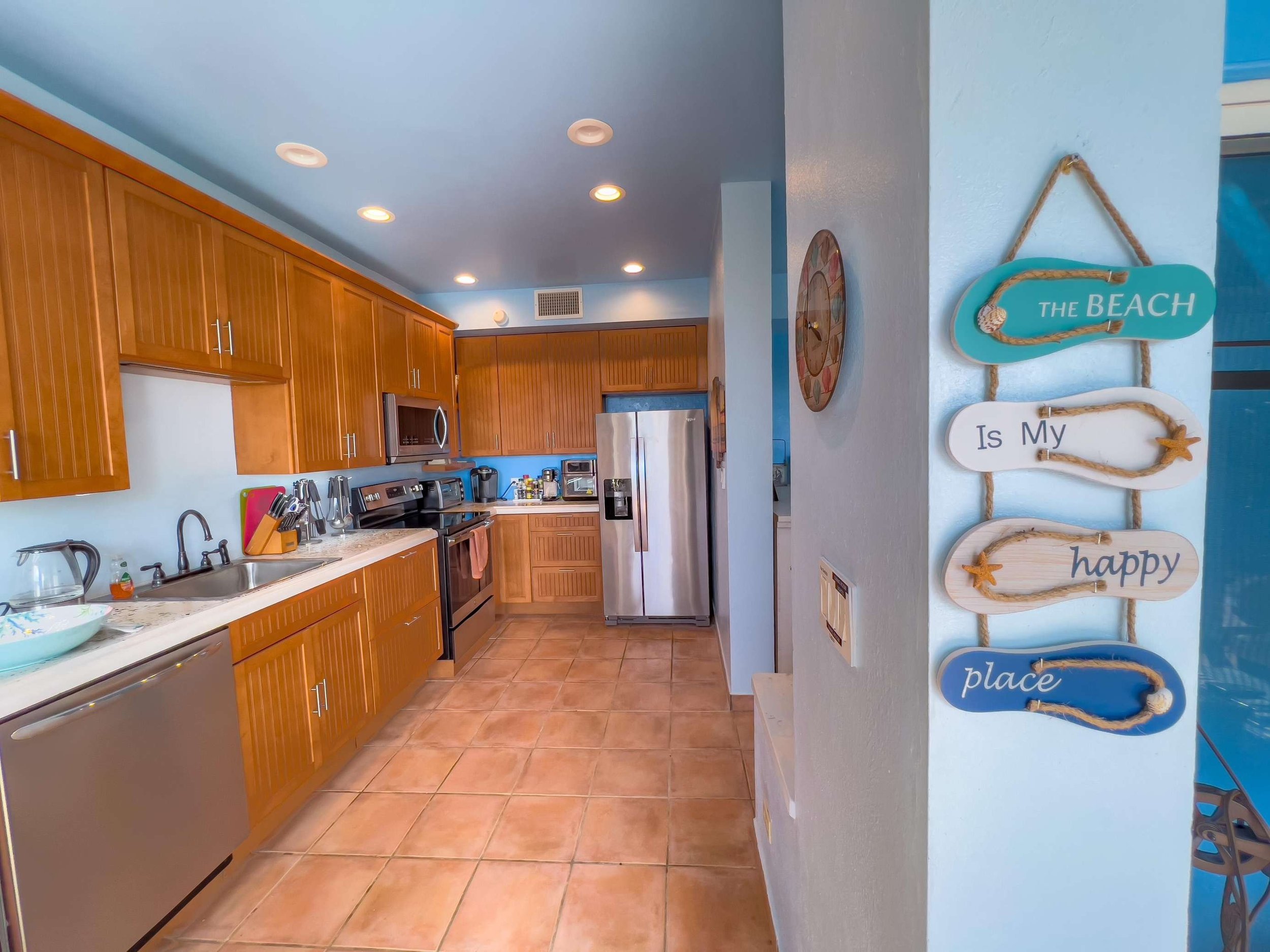 Kitchen with wooden cabinets, stainless steel appliances, and beige tile flooring, decorated with a beach-themed sign that reads 'the beach is my happy place' hanging on a white wall.
