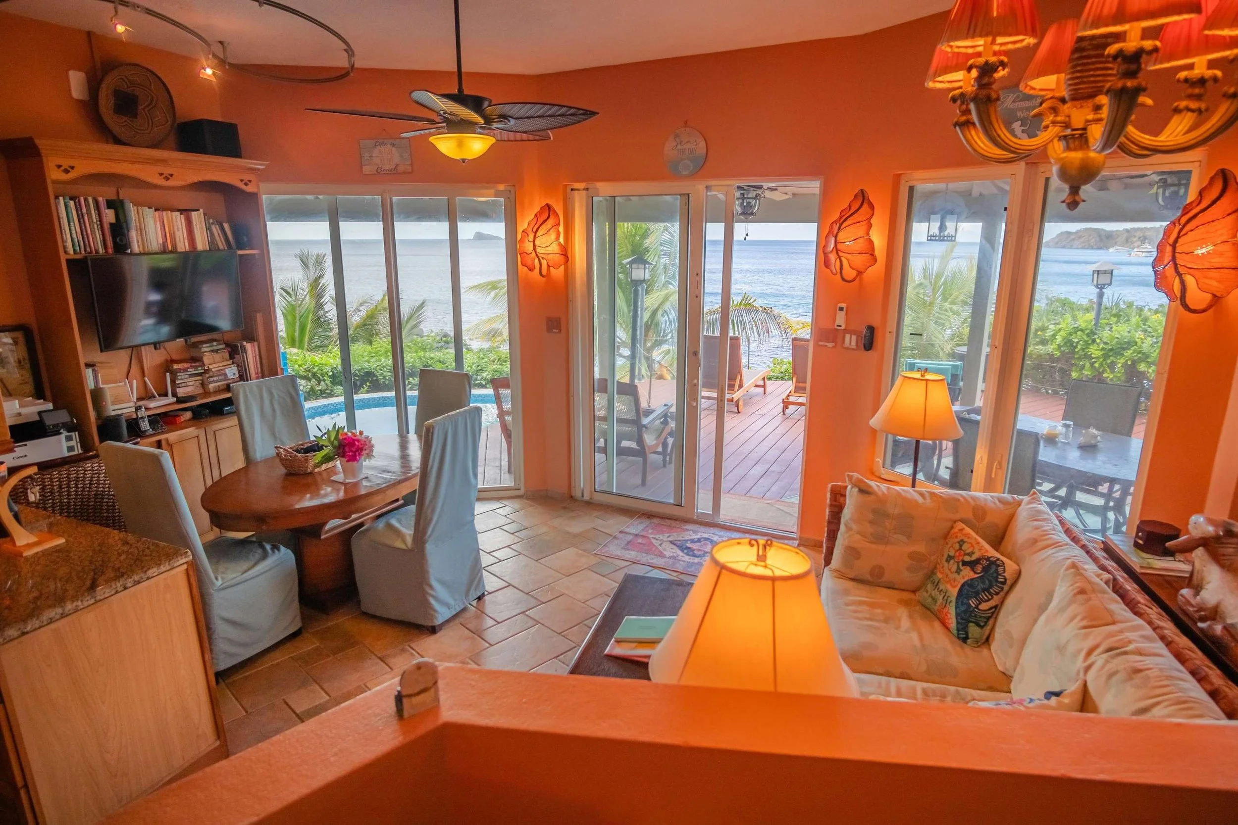 Living room with sliding glass doors leading to a patio and ocean view, featuring a dining table with chairs, a sofa, lamps, and tropical decor.