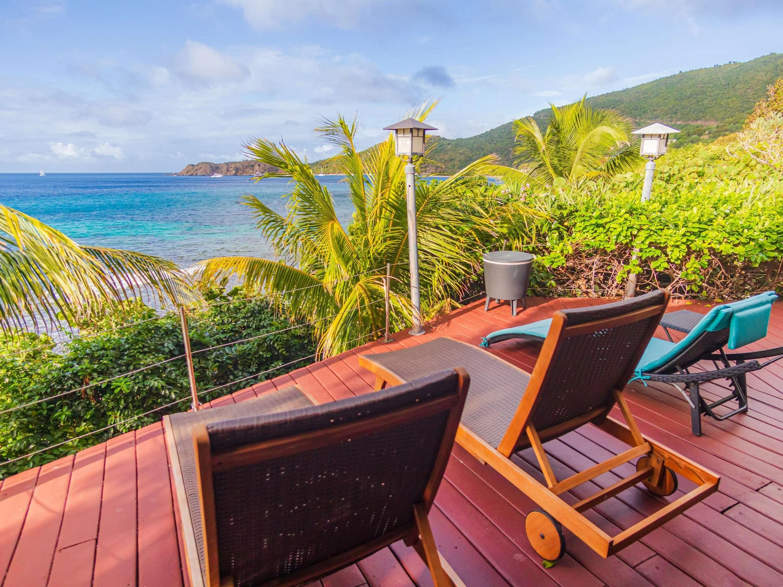 Beachside patio with lounge chairs overlooking the ocean, palm trees, and hills in the background.