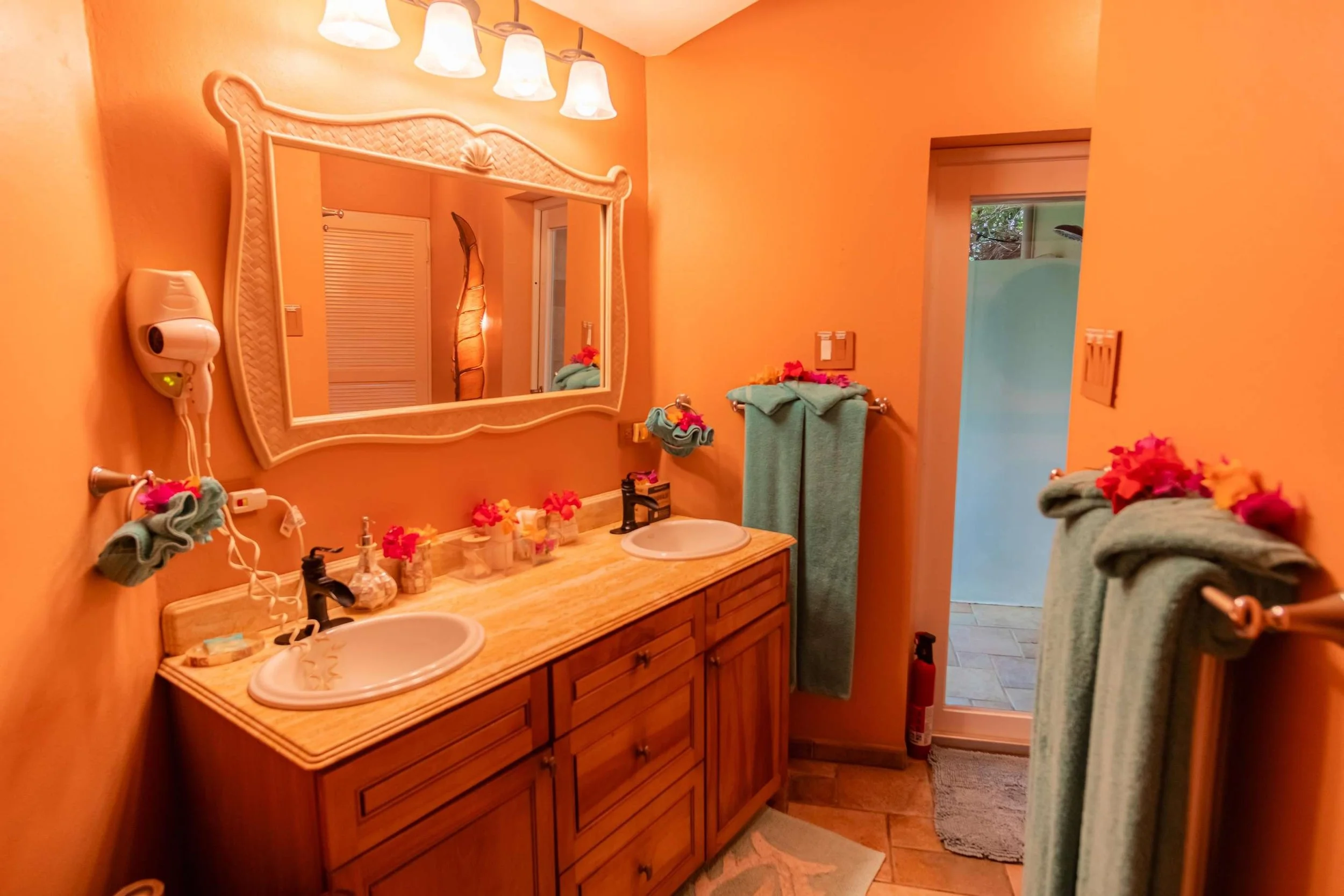 Bathroom with peach-colored walls, wooden vanity with double sinks, large mirror, light fixtures above, towels with floral decorations, and a view to an outdoor shower area.