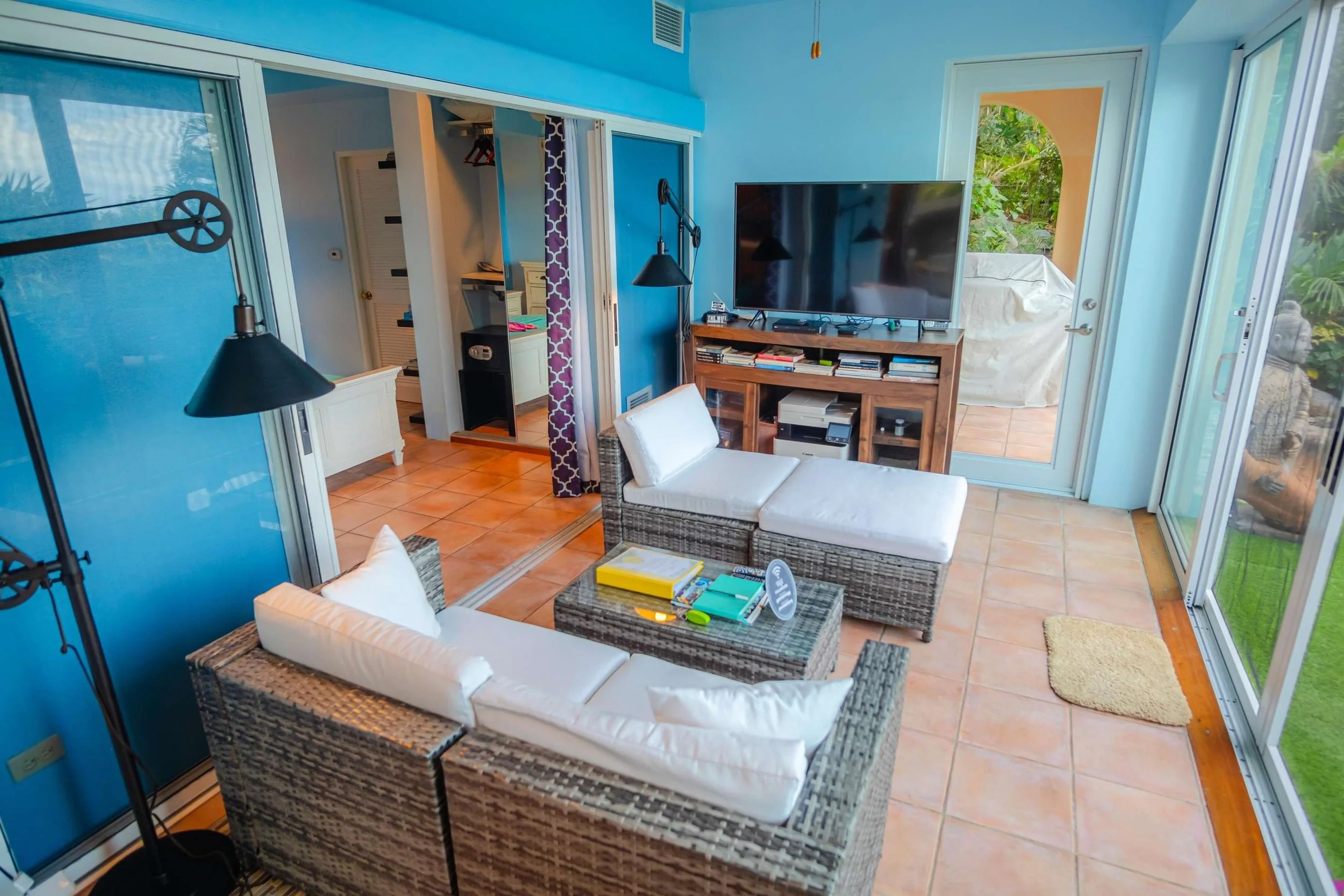 Living room with wicker furniture, a large flat-screen TV, sliding glass doors leading to a patio, and a tiled floor.