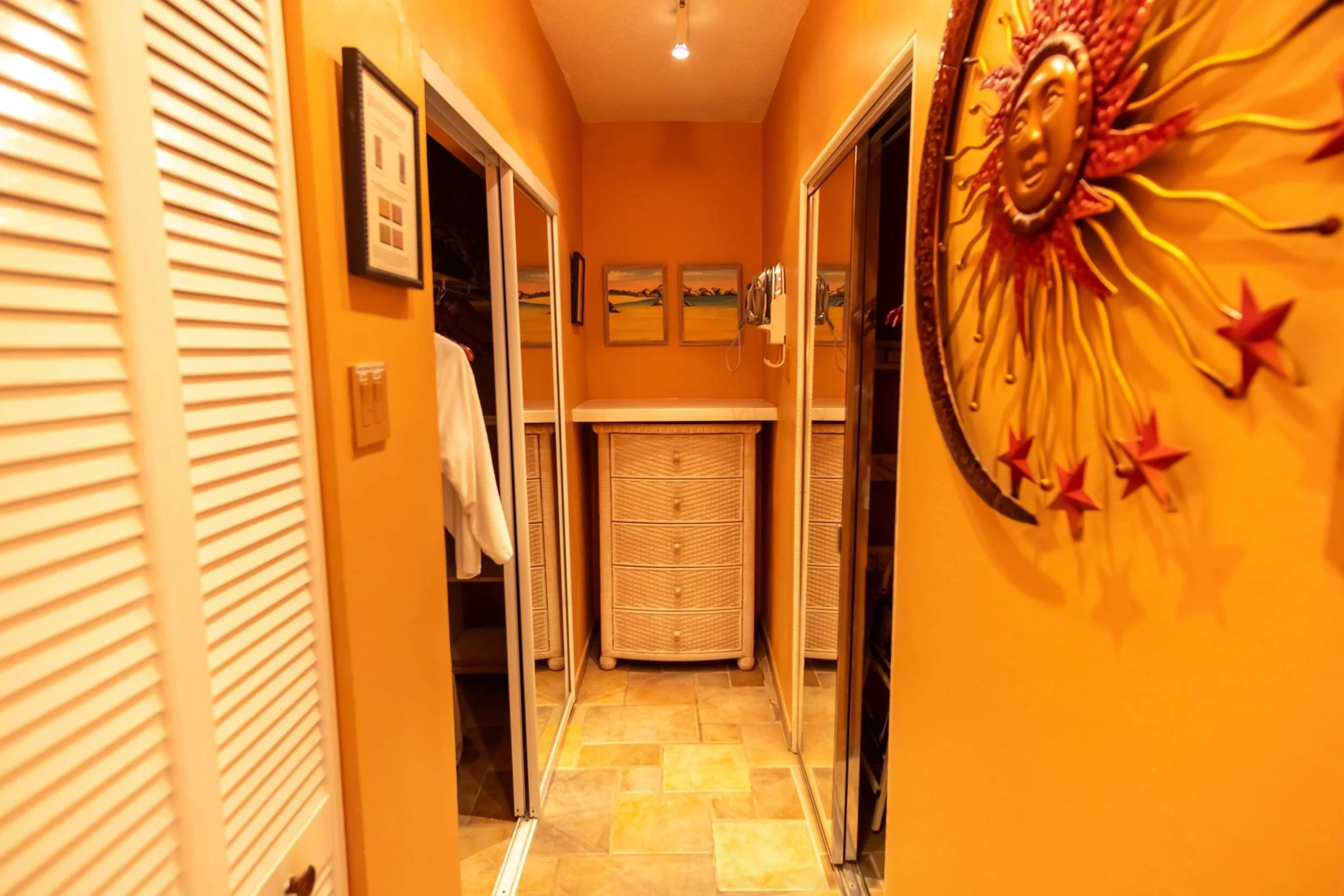 A narrow hallway with orange walls, a tiled floor, and various wall decorations including a large sun face and landscape paintings. There are mirrored closet doors on either side and a small wicker dresser at the end.