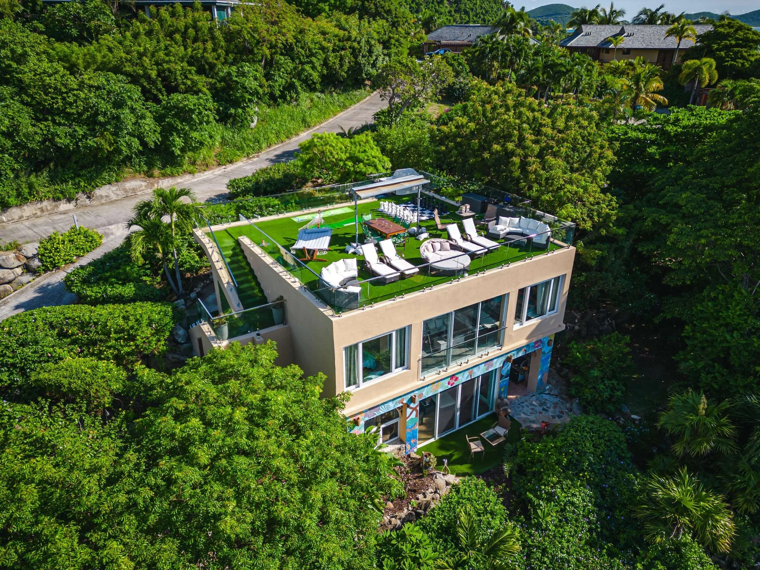 Aerial view of a modern house with a rooftop lounge area featuring outdoor furniture, lounge chairs, and a bright green artificial grass surface, surrounded by lush green trees and tropical plants.