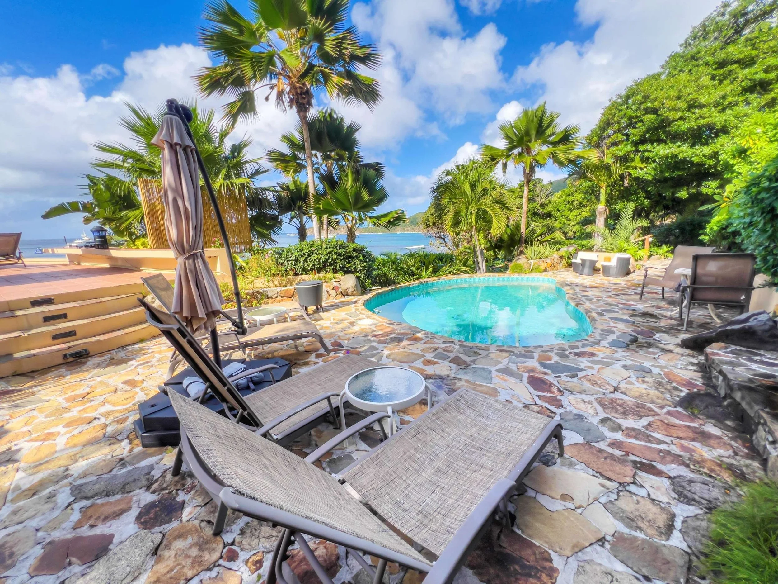 A tropical poolside scene with lounge chairs, an umbrella, lush green palm trees, and a view of the ocean in the background under a partly cloudy sky.