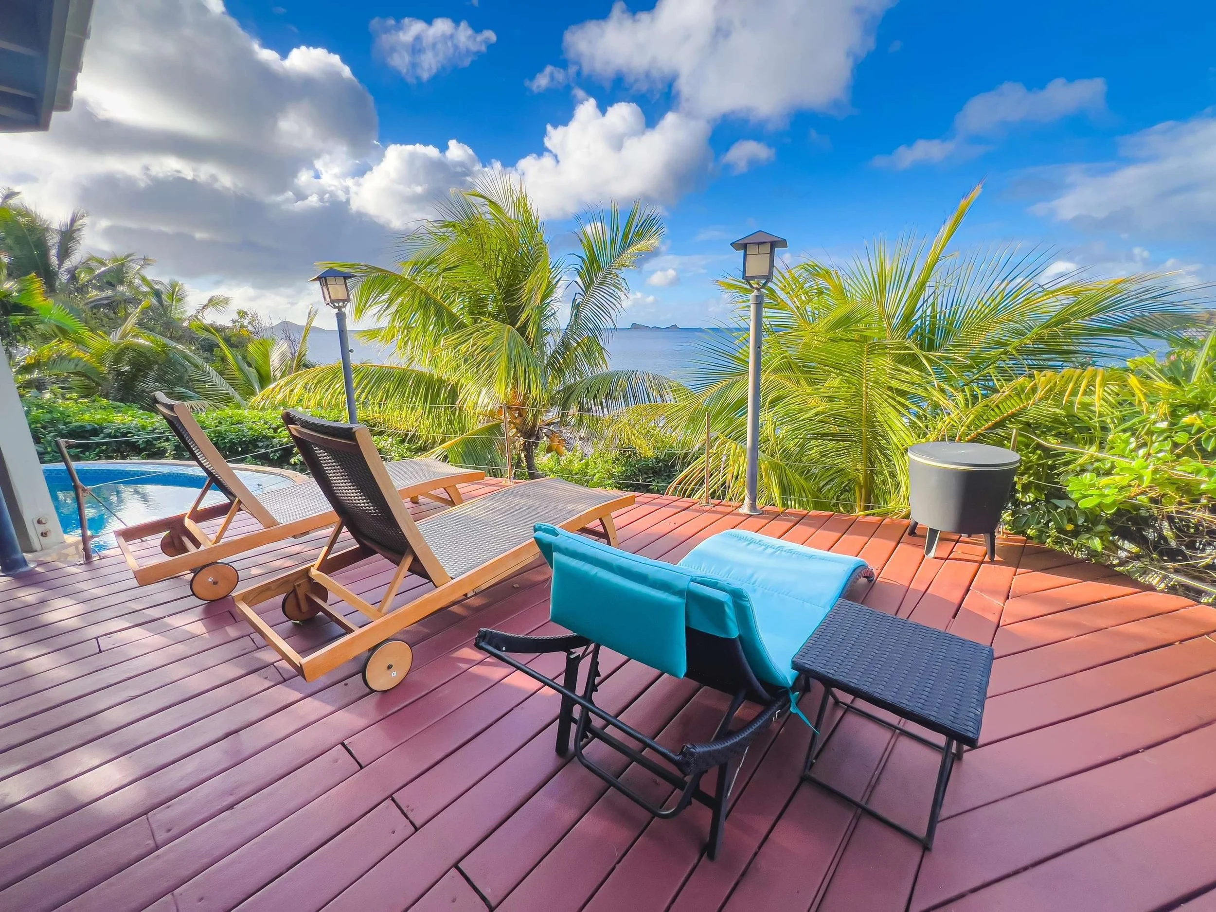 A wooden deck with two lounge chairs, a black outdoor chair with a blue towel, a small black table, and a gray outdoor stove on a reddish-brown deck, overlooking lush green palm trees and the ocean under a partly cloudy sky.