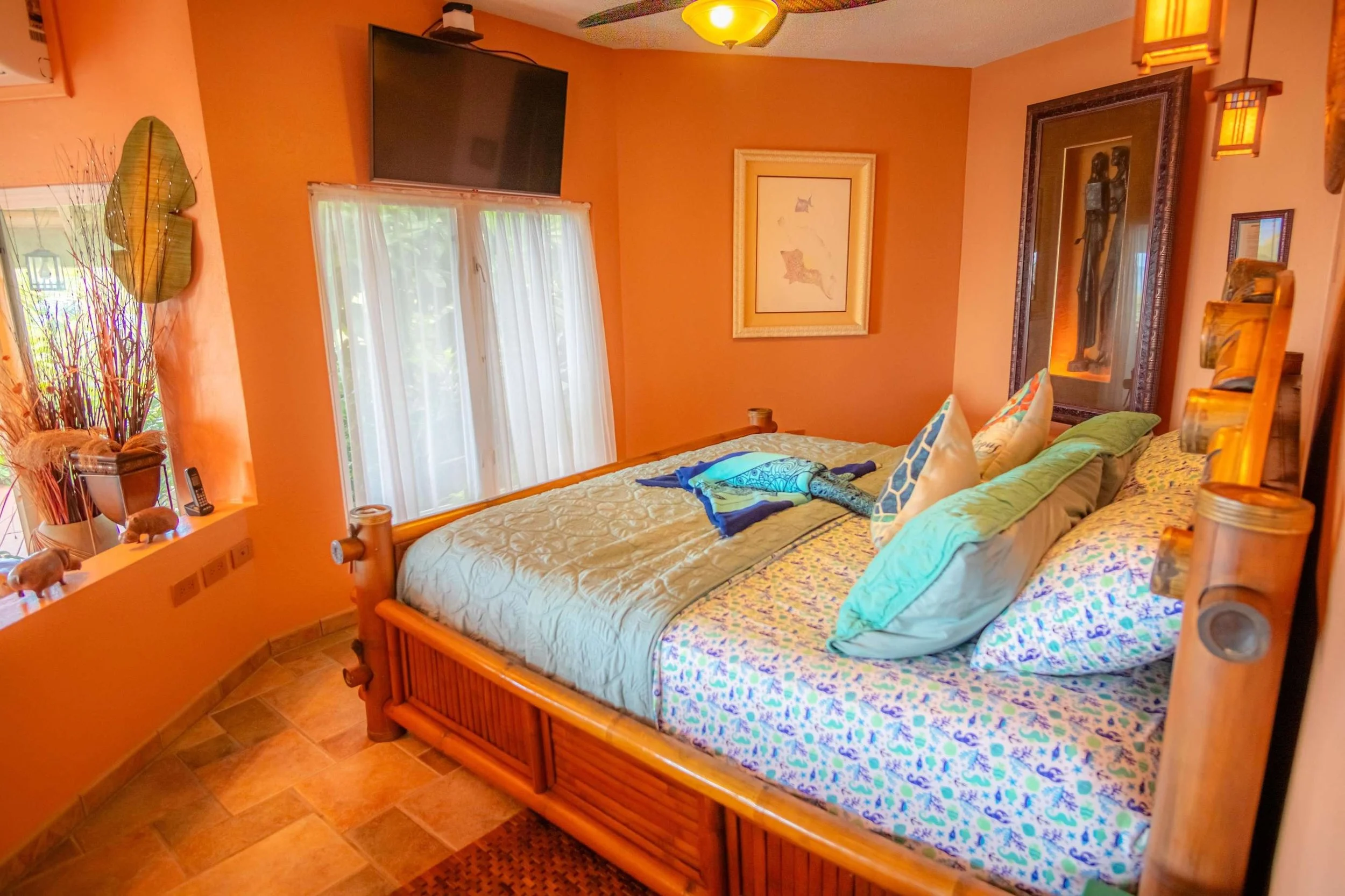 A cozy bedroom with orange walls, bamboo bed frame, and a colorful floral bedspread. Decor includes framed art, wall hanging, a flat-screen TV, and tropical accents like large leaves and decorative animals by the window.