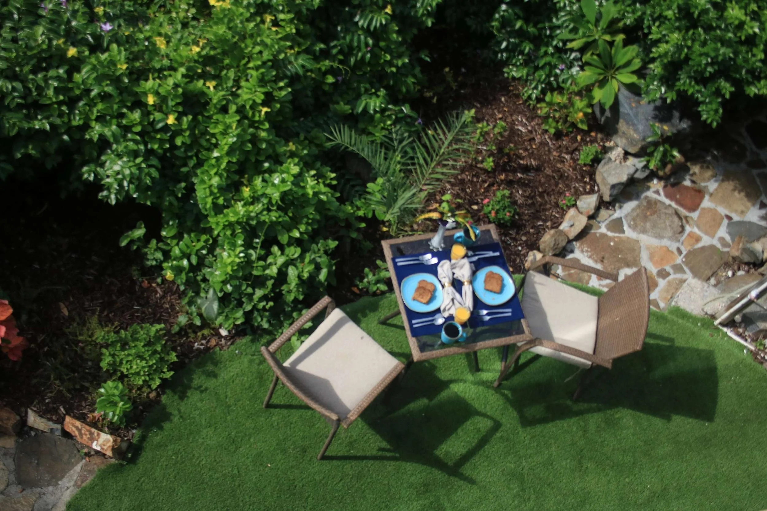 An outdoor dining area with two chairs and a small table set for breakfast, featuring two plates with toast, cups, and utensils, surrounded by green bushes and a stone pathway.