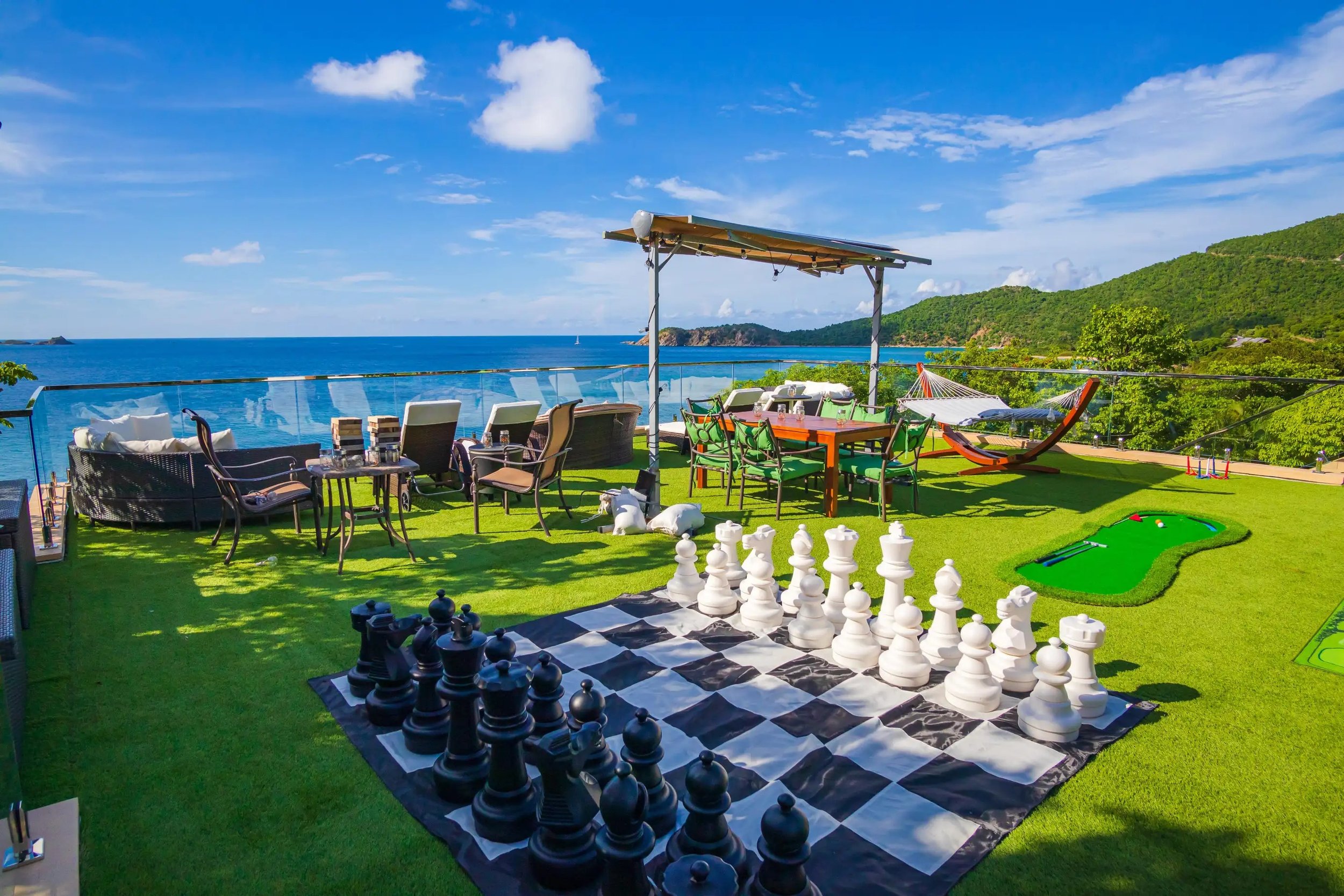 Outdoor terrace with seating, a large inlaid chessboard, a miniature golf putting green, a hammock, and a view of the ocean and green hills in the background.