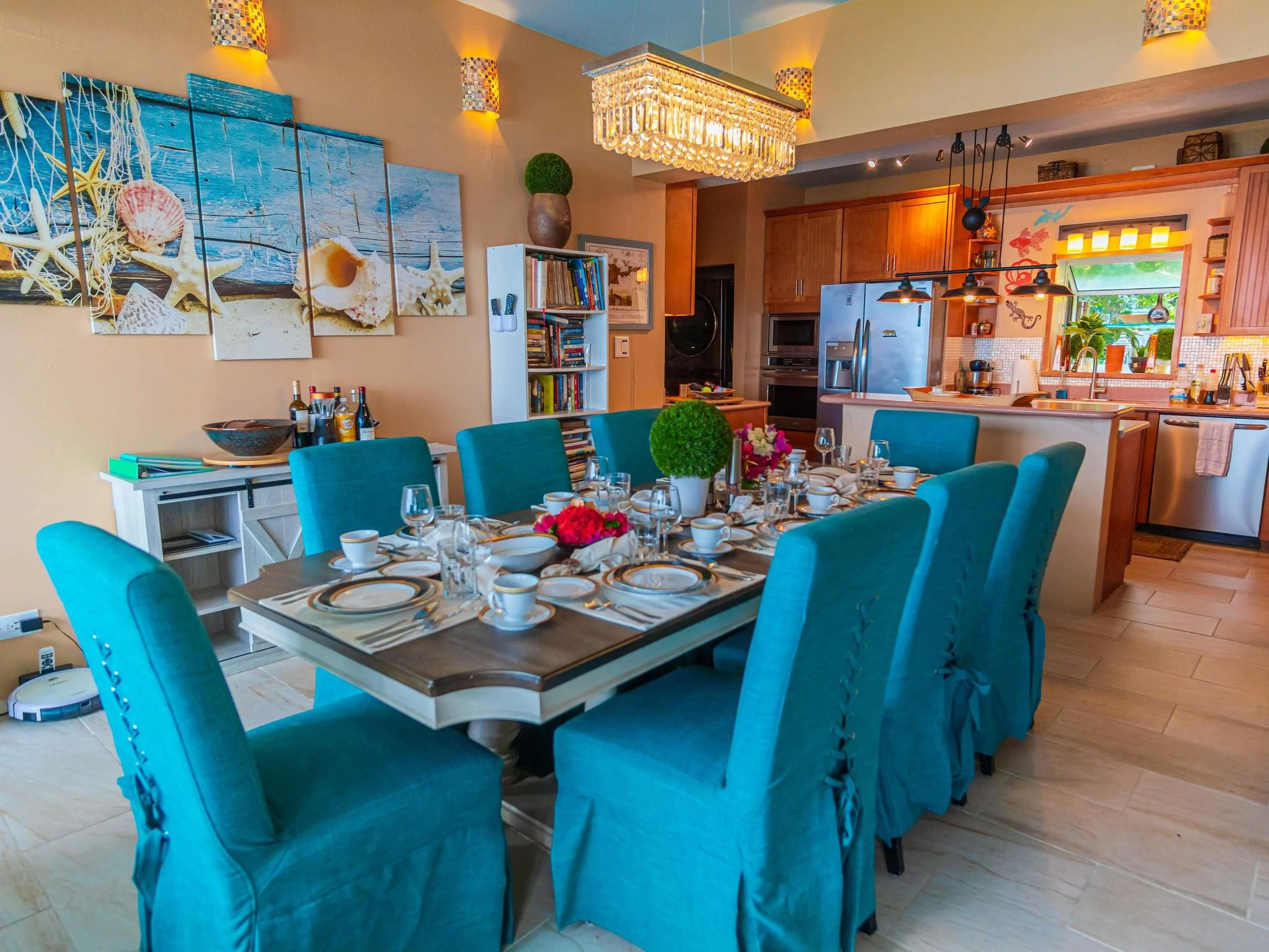 A dining table set for a meal with blue upholstered chairs, white dinnerware, glasses, and floral centerpieces, in a brightly decorated kitchen and dining area with beach-themed artwork and warm lighting.