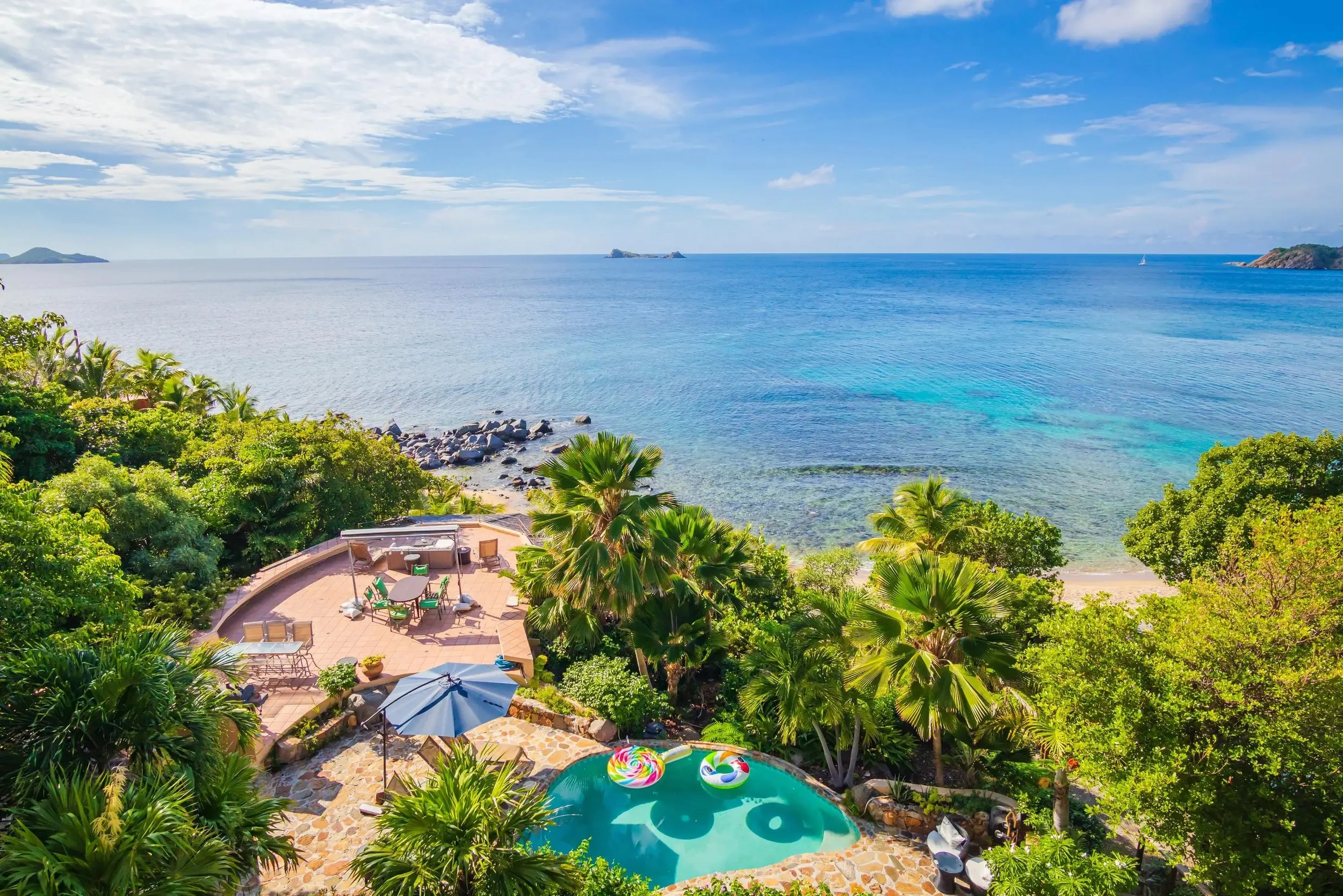A scenic view of a tropical seaside property with a swimming pool, lush green palm trees, and a patio with outdoor furniture, overlooking the ocean with clear blue waters and a partly cloudy sky.