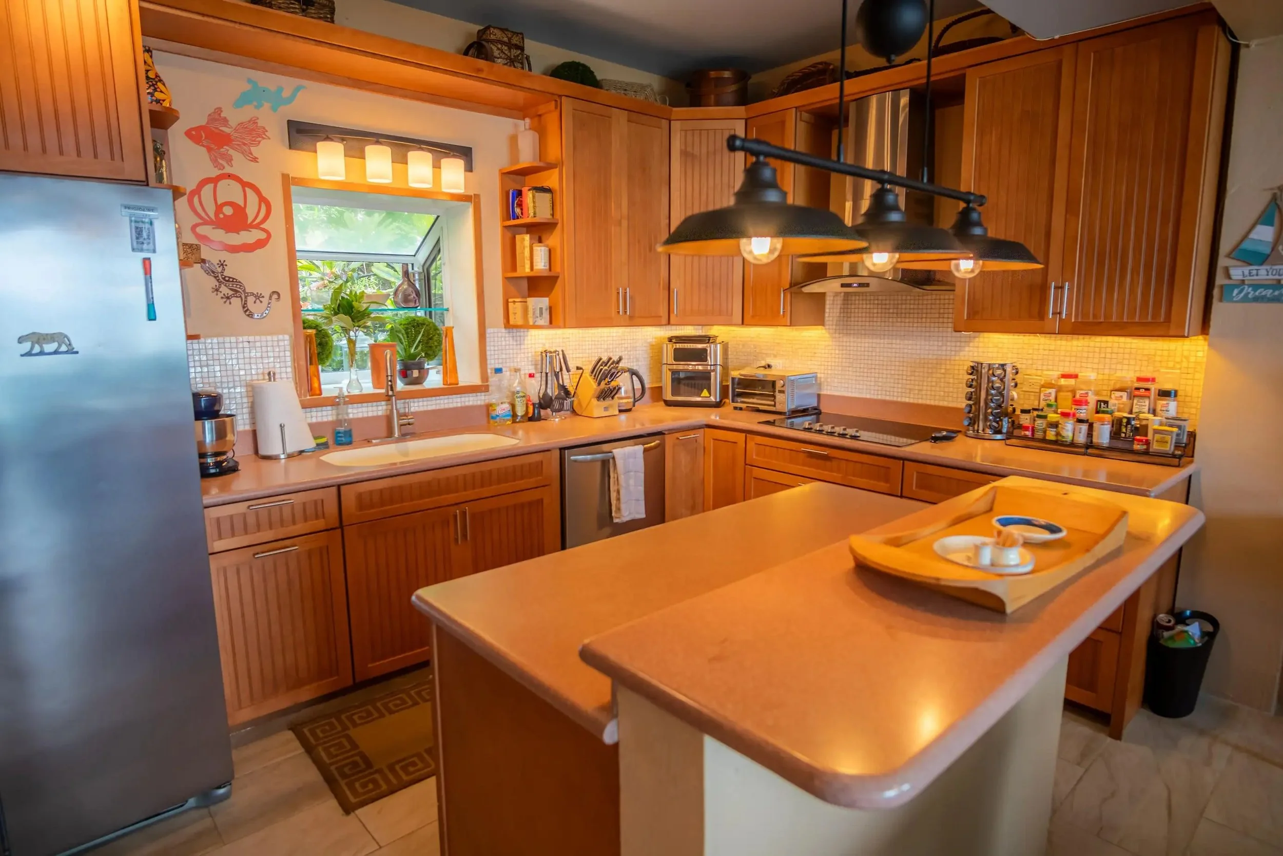 A kitchen with wooden cabinets, a small window over the sink showing greenery outside, and various appliances on the countertop. There's a kitchen island with a wooden tray and some small items. Over the island hangs a black industrial-style light fi