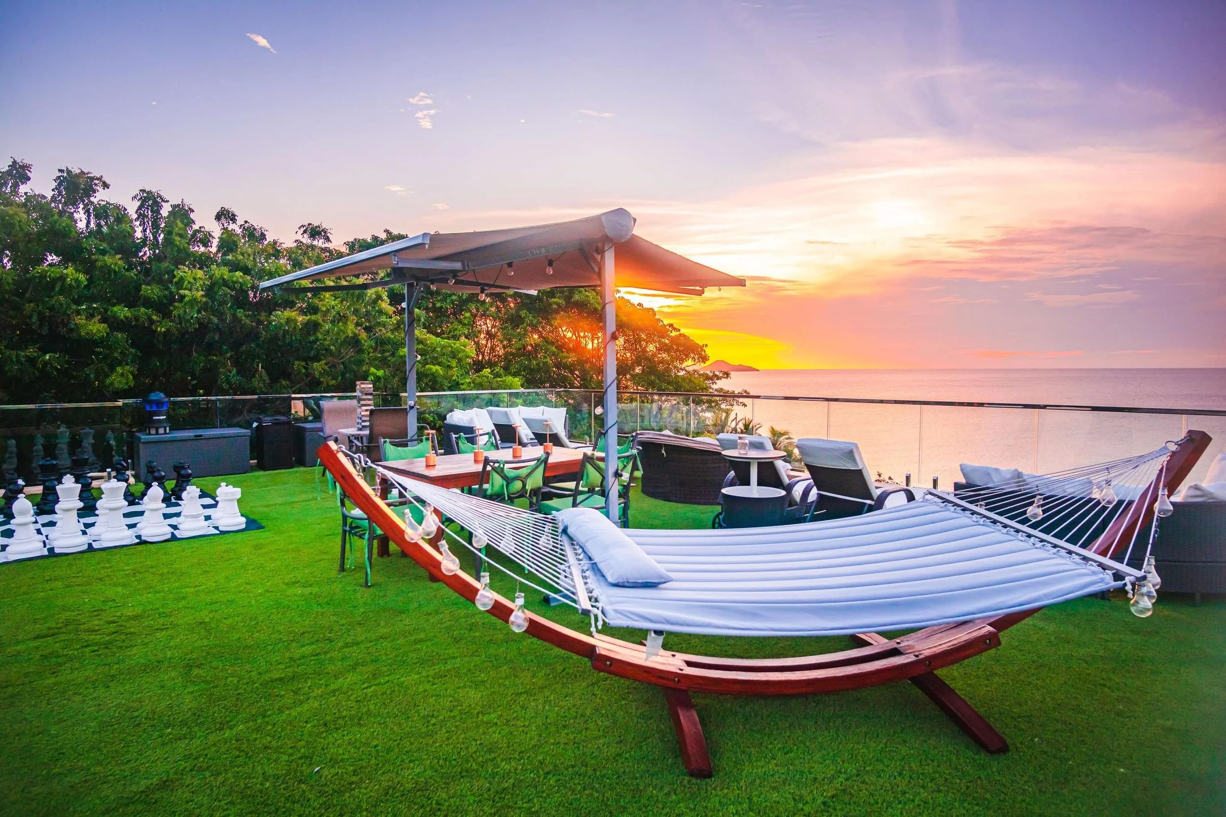 A rooftop patio overlooking the ocean at sunset with a hammock, outdoor furniture, a chess set, and lush greenery.