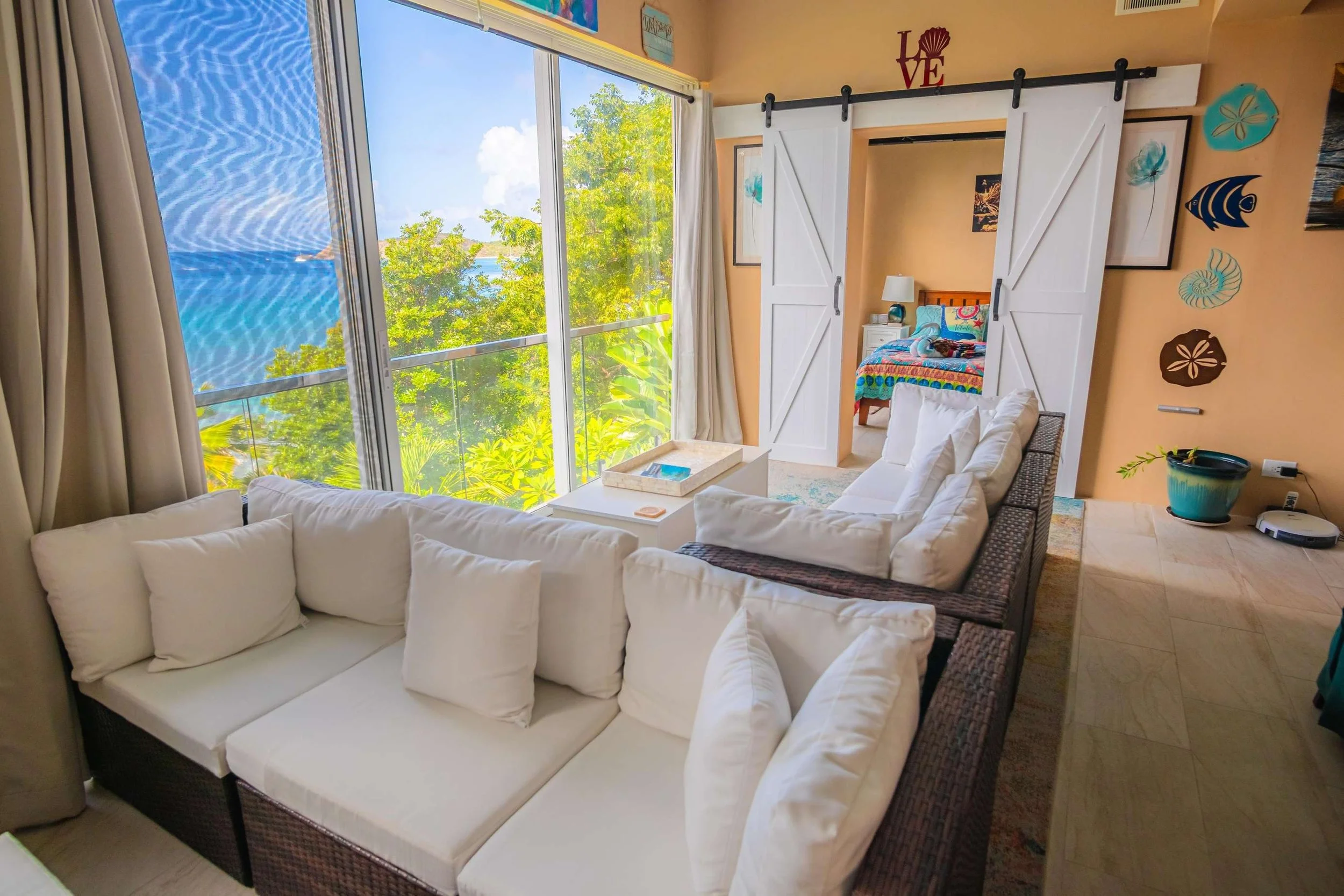 Living room with large window overlooking trees and water, white sectional sofa, and view into a bedroom with colorful bedspread, ocean scene artwork, and sunset decoration on yellow walls.