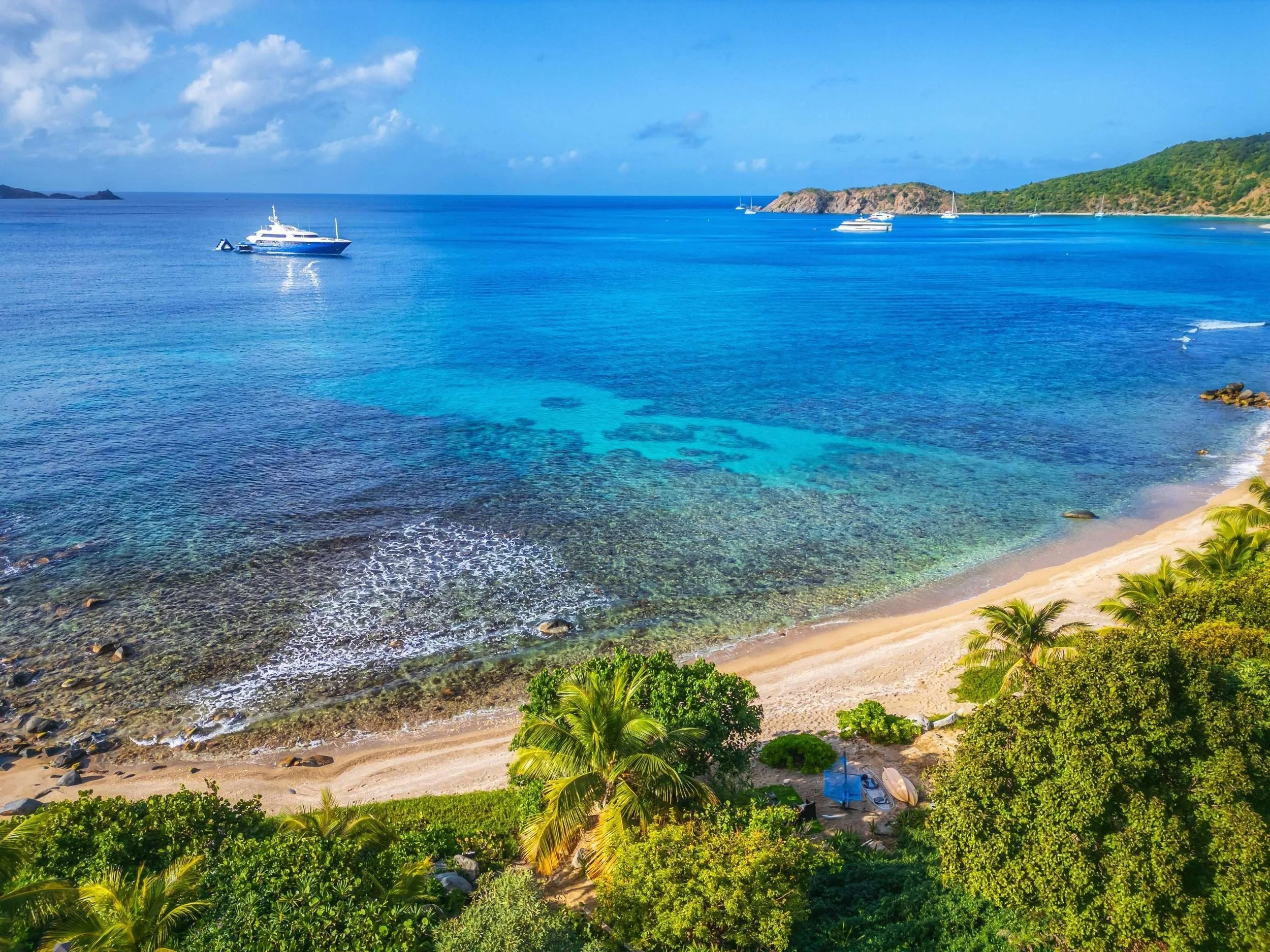 Tropical beach with clear blue water, sandy shoreline, palm trees, and boats anchored in the water under a partly cloudy sky.