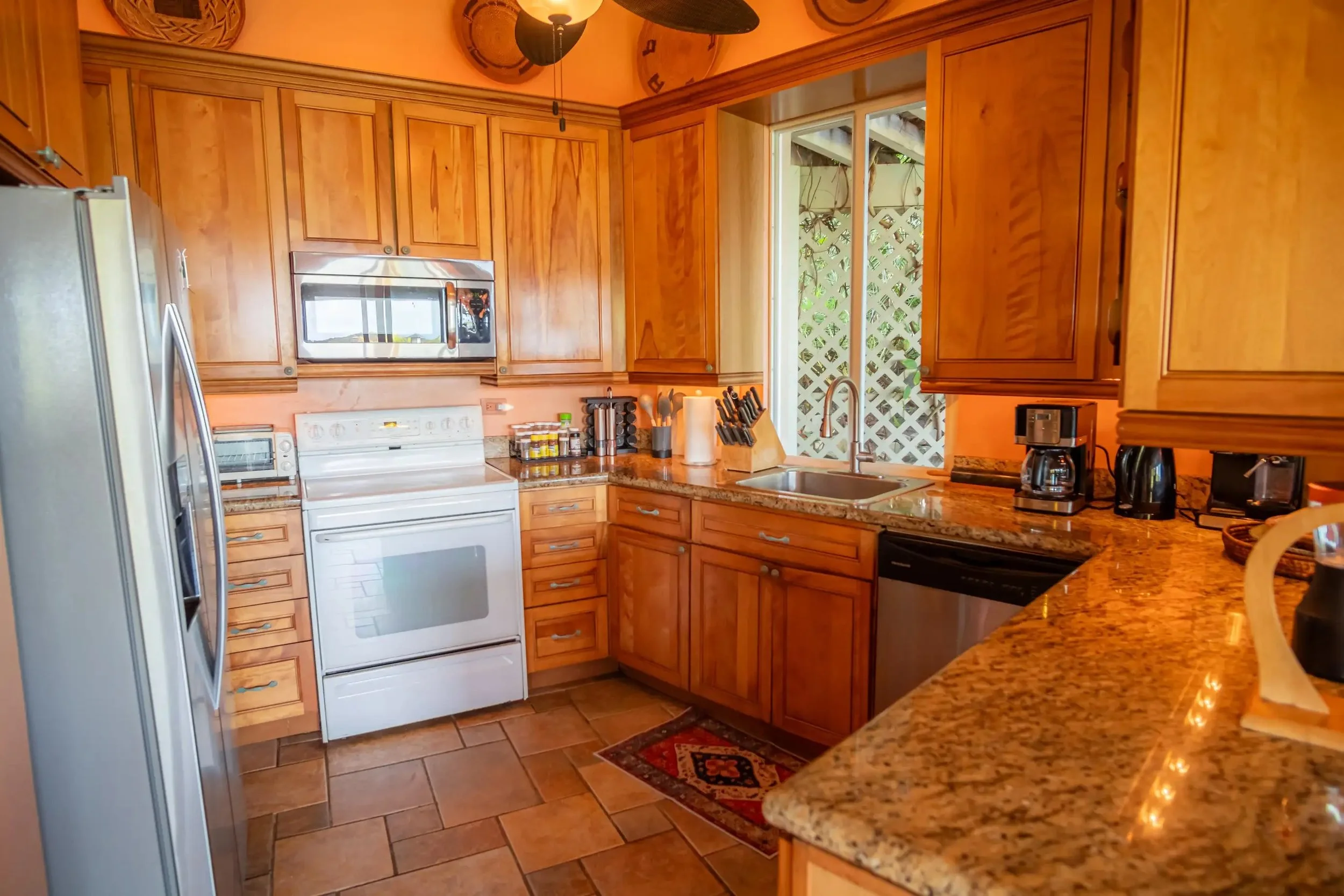 A kitchen with wooden cabinets, granite countertops, a white stove, microwave, refrigerator, coffee maker, toaster, and a double sink near a window with a lattice outside.