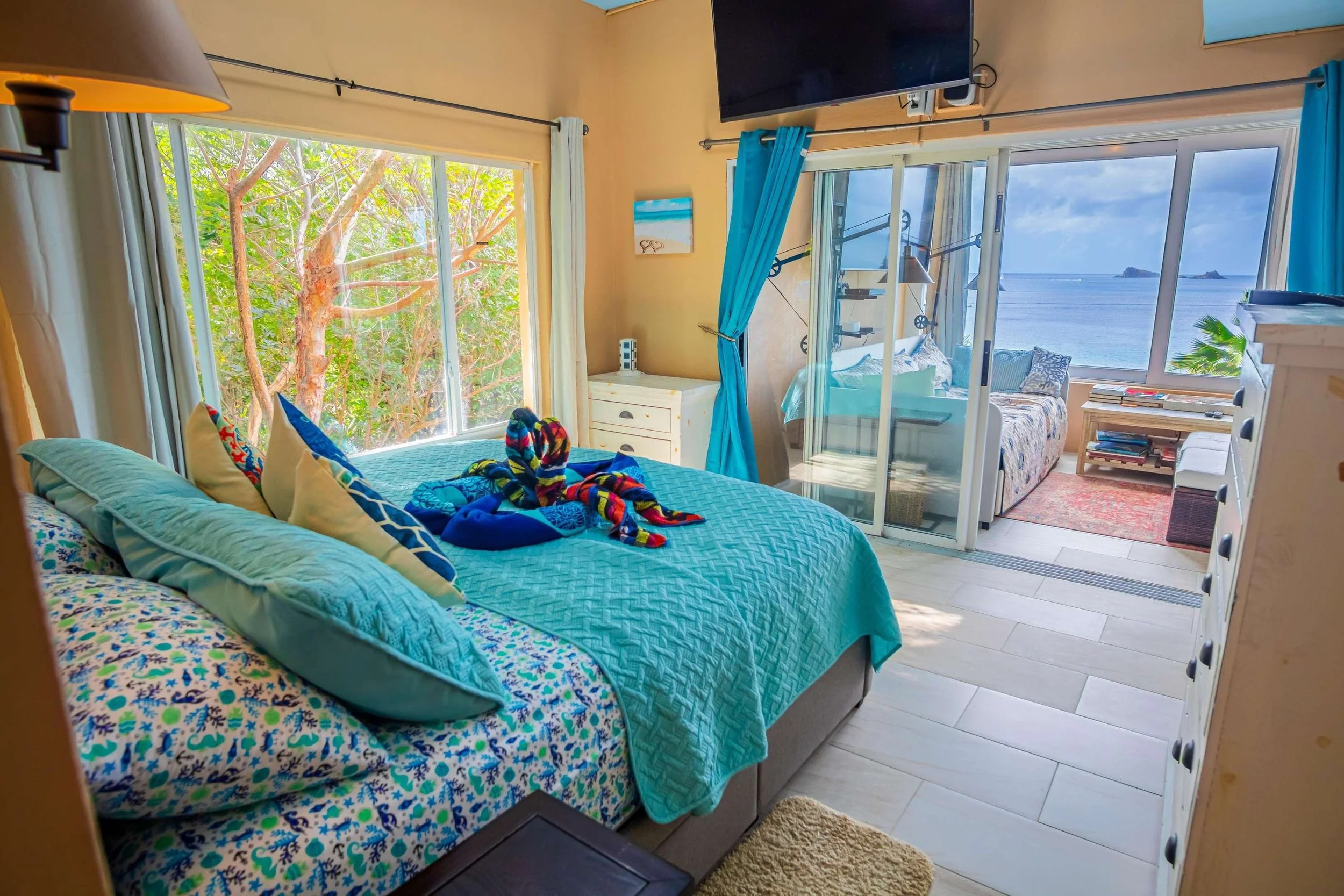 A bedroom with a large bed covered in blue and patterned bedding, decorated with colorful towel animals, next to windows showing greenery and ocean views, a sliding glass door leading to a balcony with ocean view, and a flat-screen TV mounted on the 