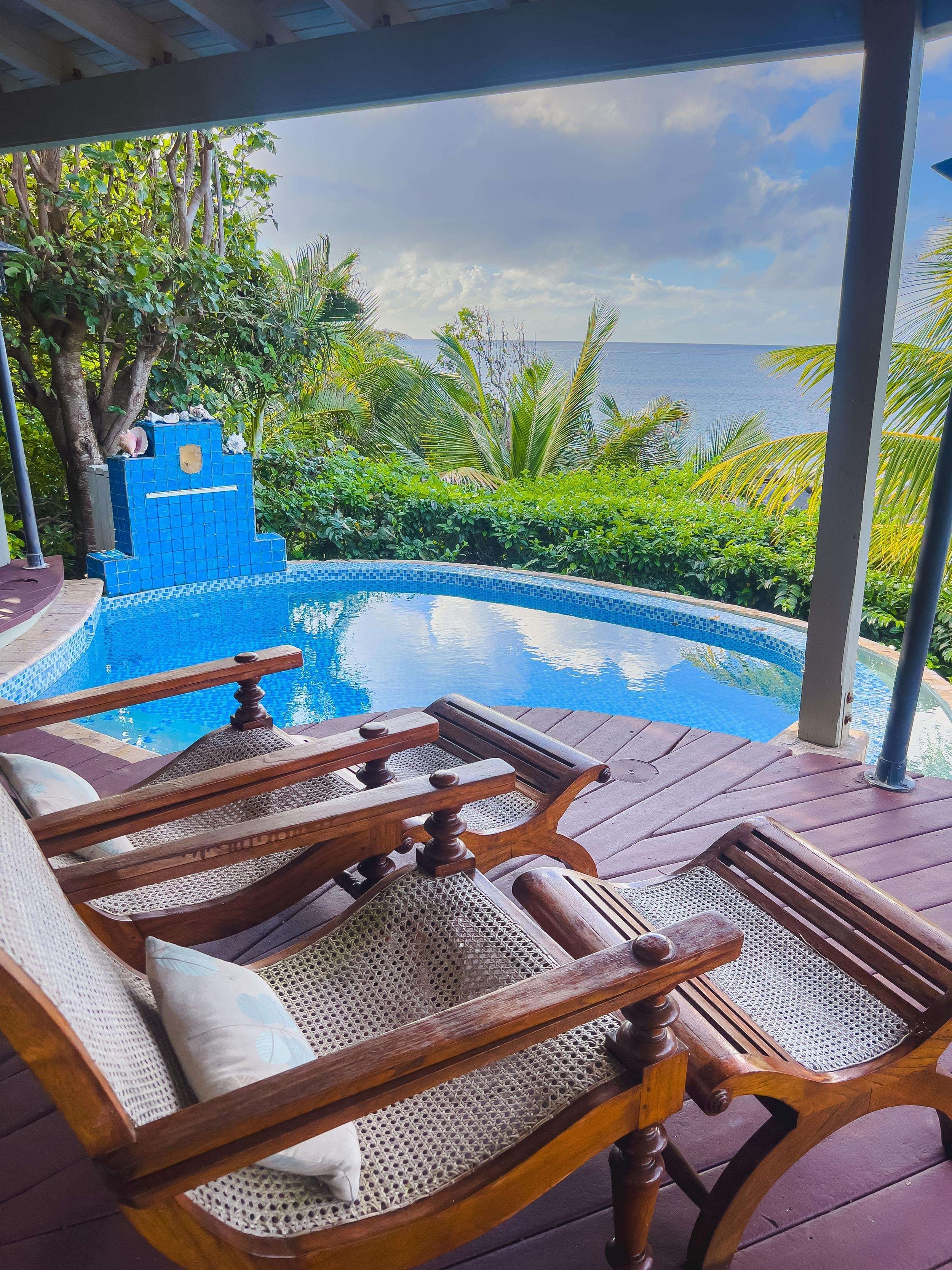A view of a pool with lounge chairs, overlooking a tropical landscape with palm trees and the ocean in the background.