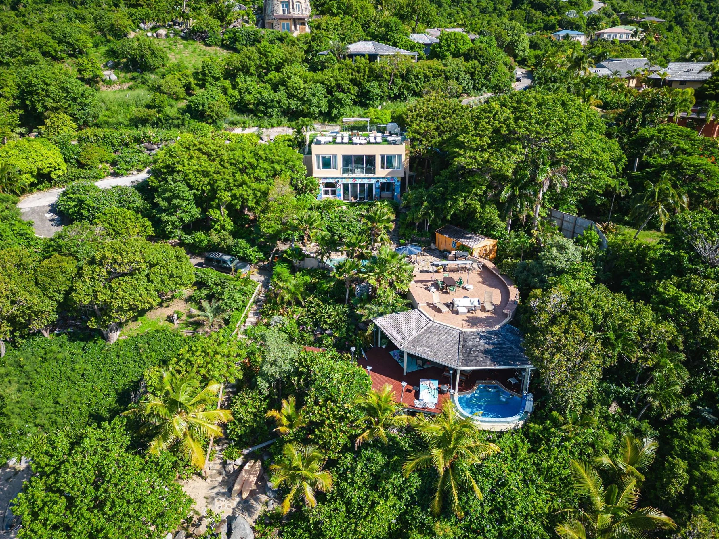 A lush, green hillside residential property featuring a modern house, a swimming pool, outdoor sitting areas, and surrounded by dense trees and tropical vegetation.