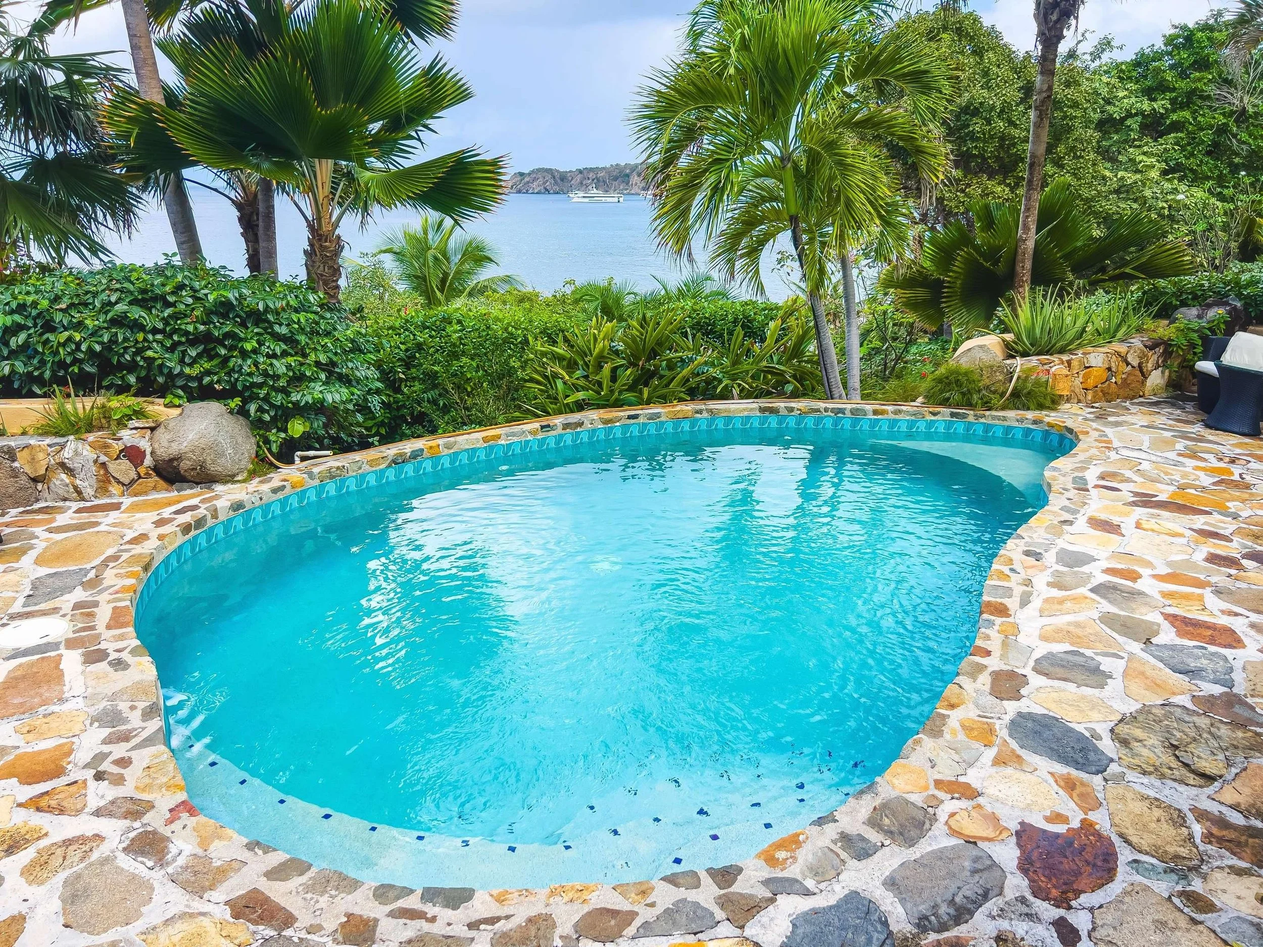A turquoise swimming pool surrounded by a stone patio and lush green tropical plants, with a view of a body of water and trees in the background.