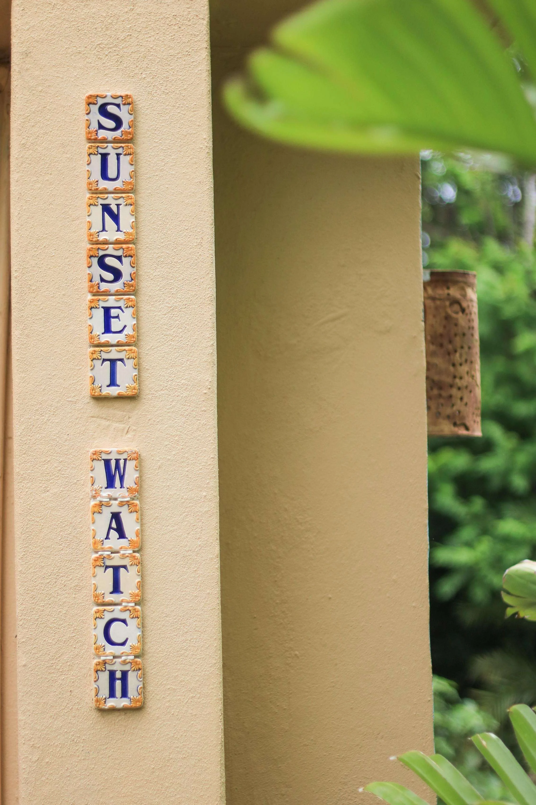 Ceramic tiles on a wall spell out "Sunset Watch" in vertical orientation with a beige stucco wall and green foliage in the background.