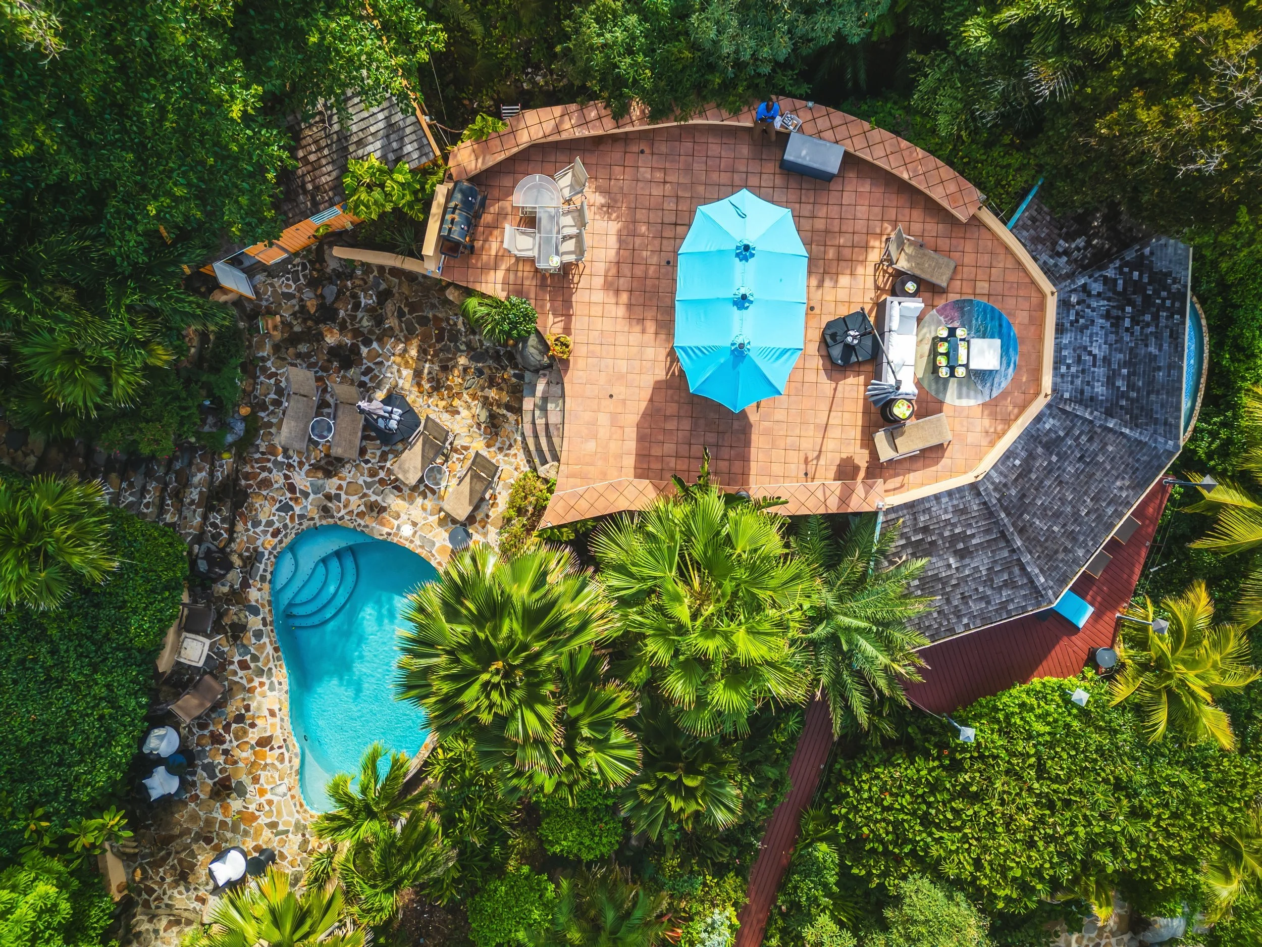 Aerial view of a backyard with a swimming pool, surrounded by lush green trees and plants. There is a tiled patio area with outdoor furniture, including a table with an umbrella, lounge chairs, and a barbecue grill.