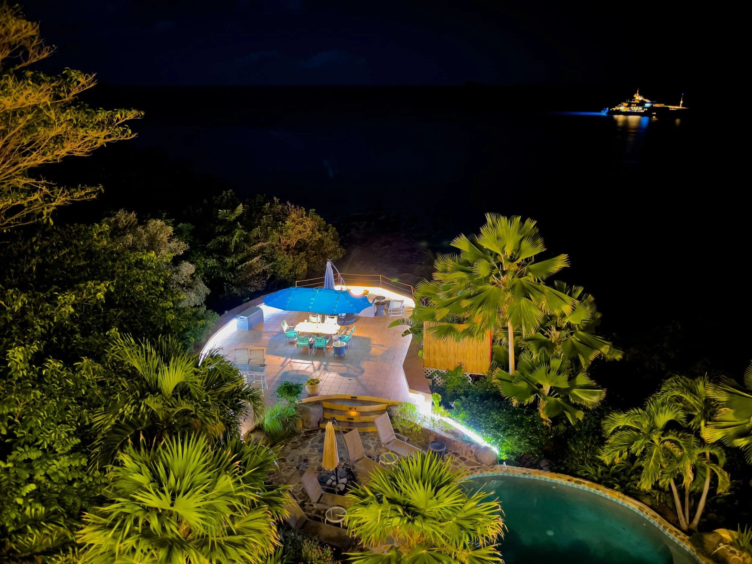 Night view of a decorated outdoor patio with a large blue umbrella, surrounded by tropical plants, overlooking the ocean with a yacht in the distance.