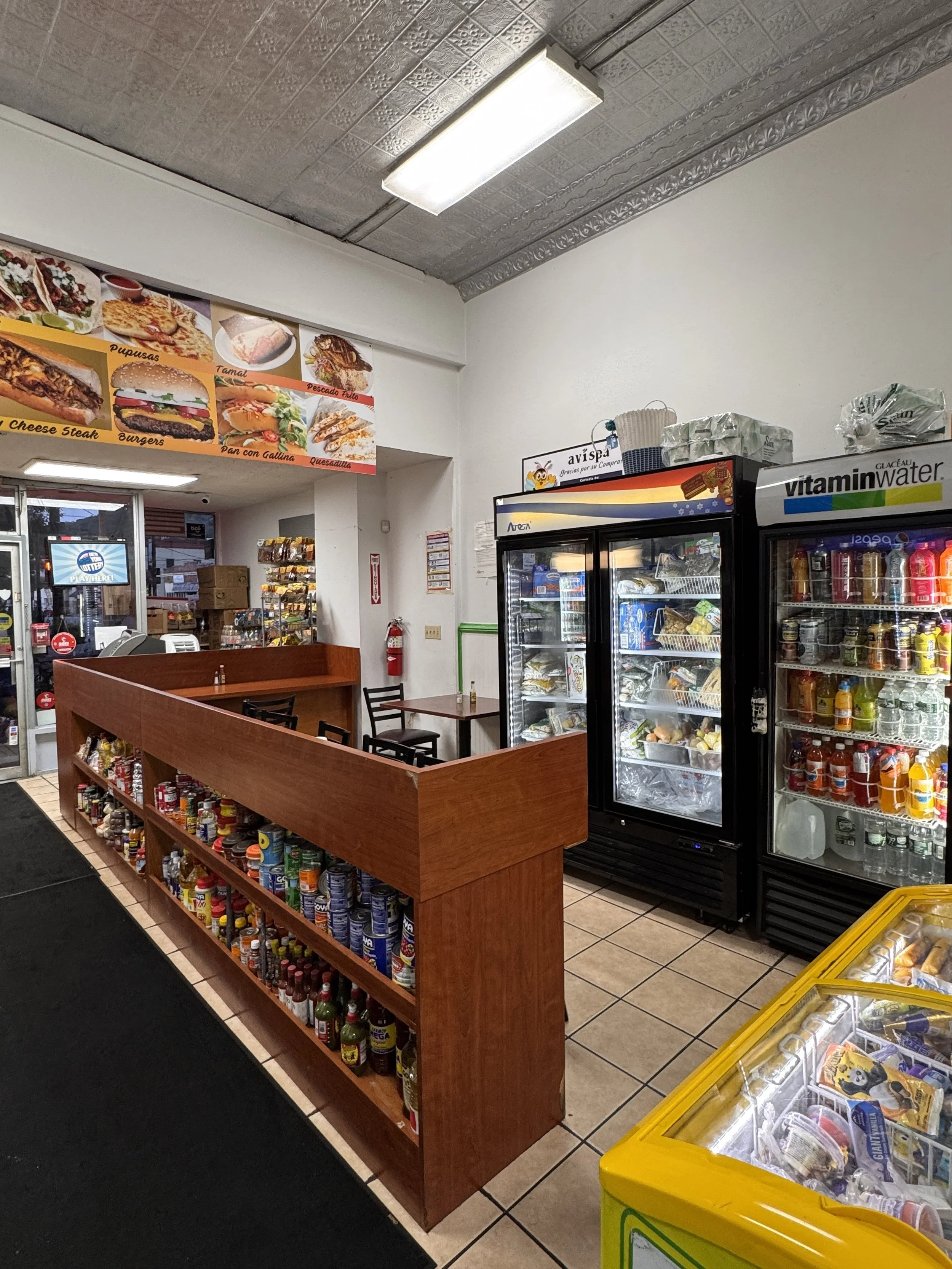 Interior of a small convenience store or fast food restaurant showing refrigerator units, shelves with snacks and drinks, and a black and yellow ice chest. There are chairs and a small table near the wall, with menu images of food items above the ent