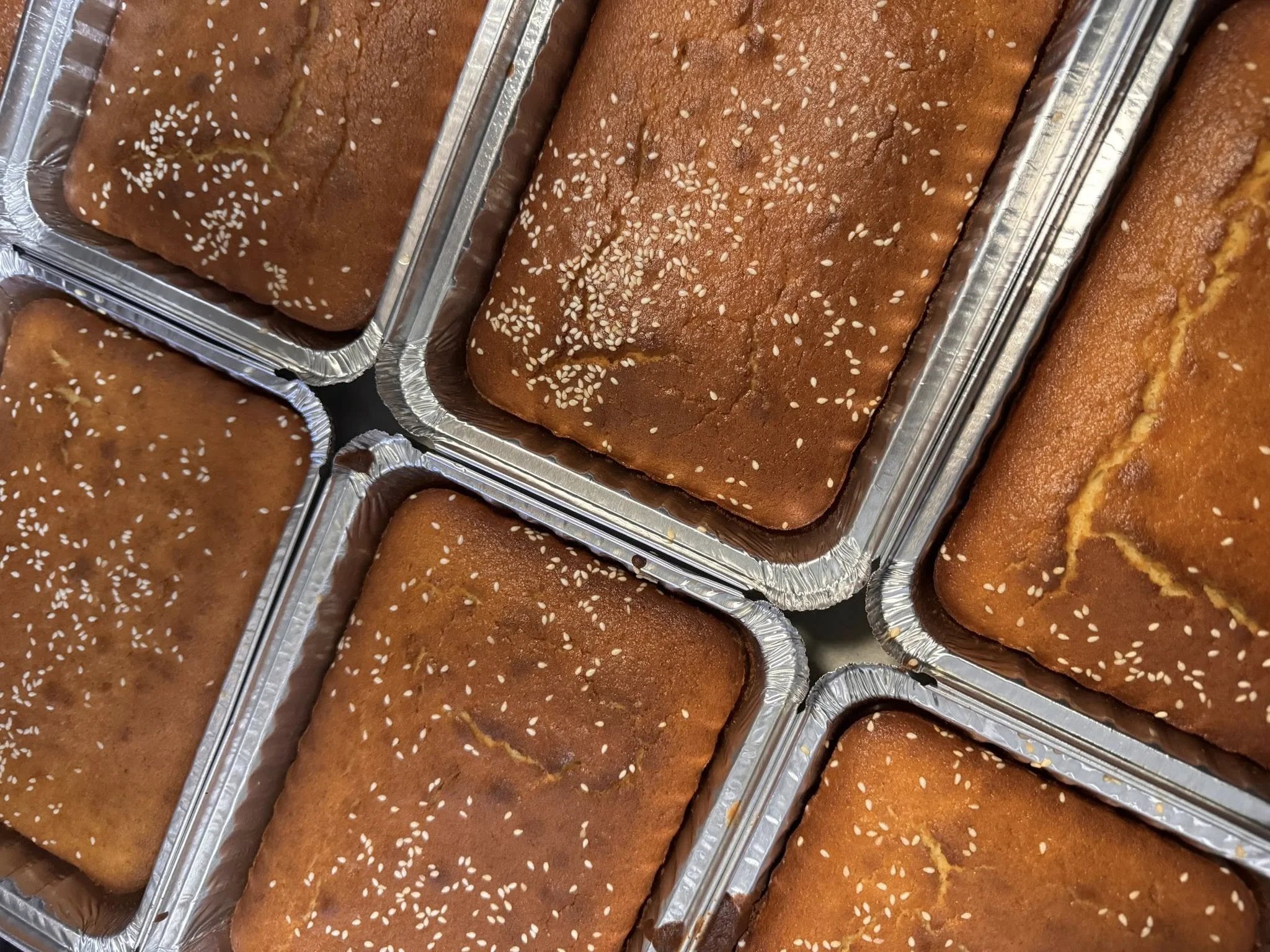 Six freshly baked bread in aluminum foil pans, sprinkled with sesame seeds.